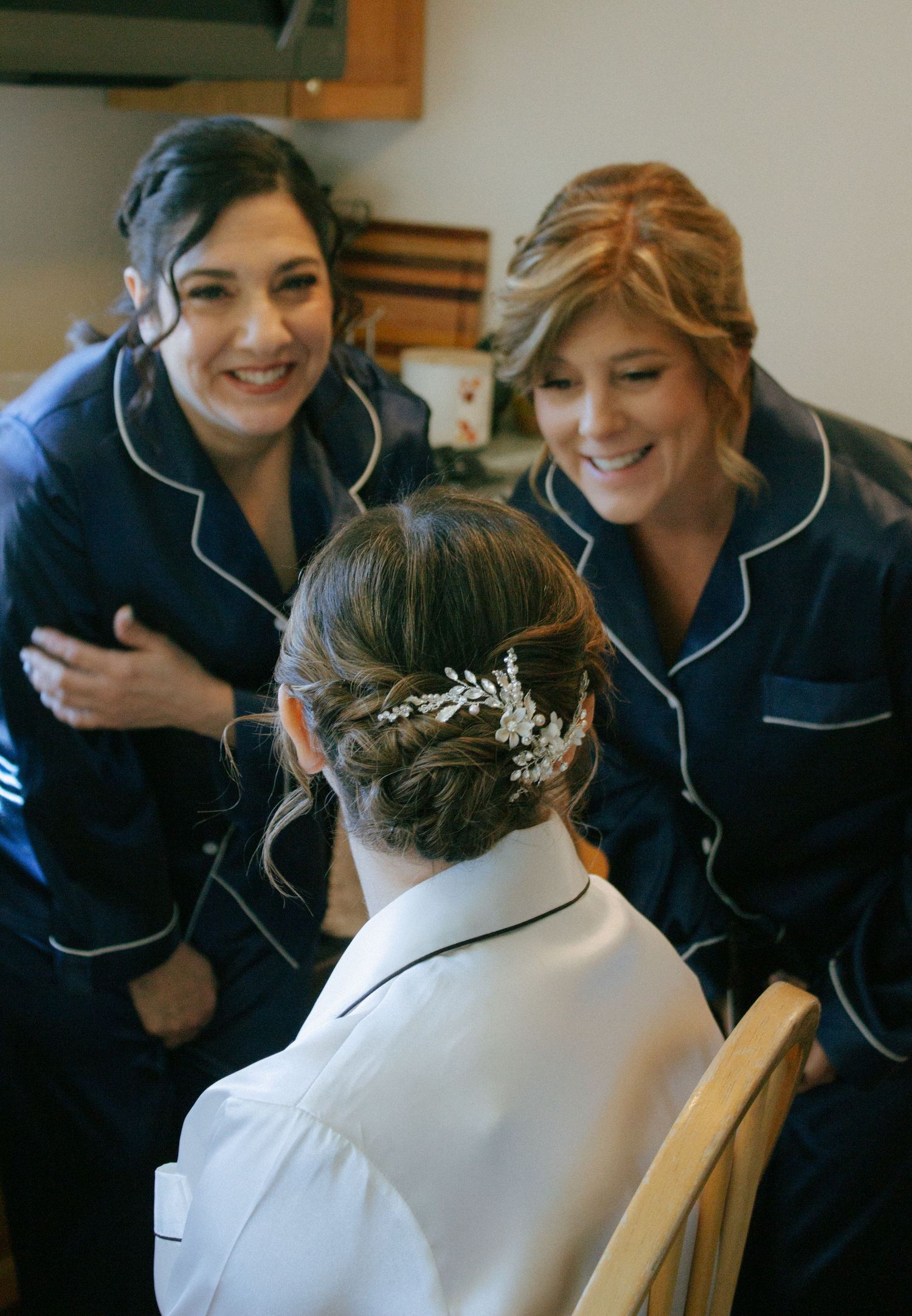 Two bridesmaids in navy pajamas smile warmly at a bride with an elegant braided updo hair accessory.