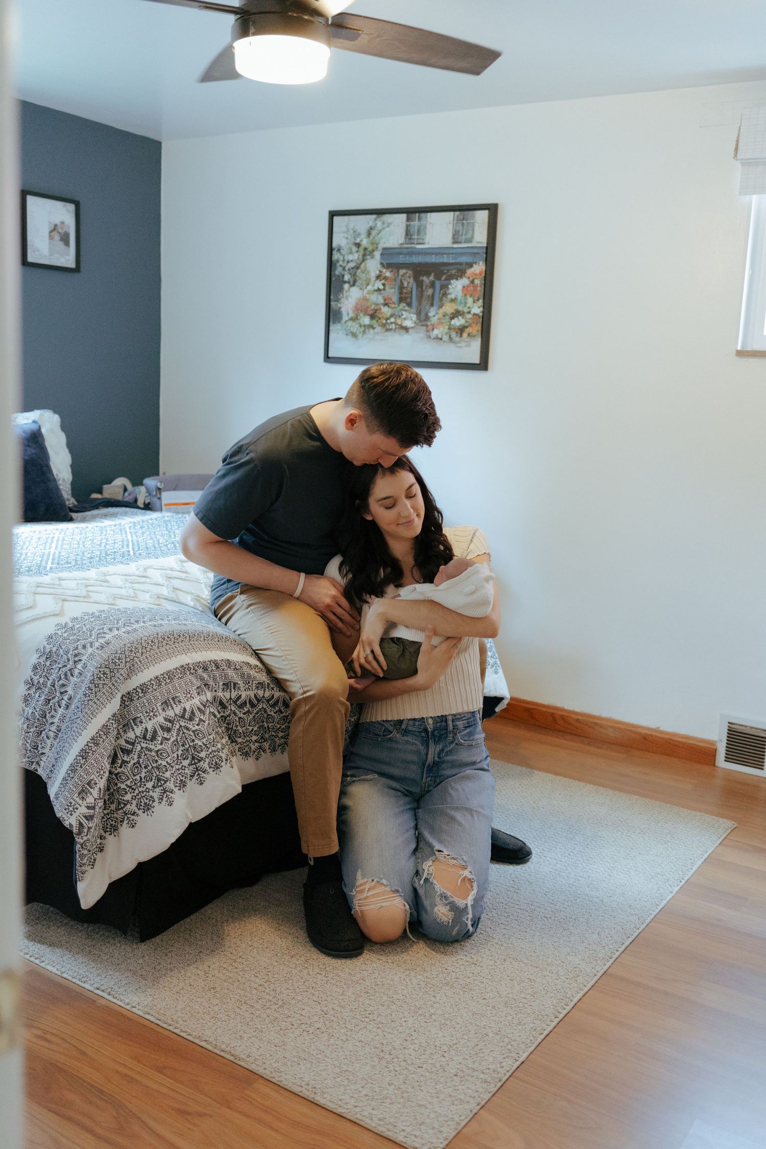 Young couple kneeling by bed holding newborn baby in cozy bedroom with hardwood floors.