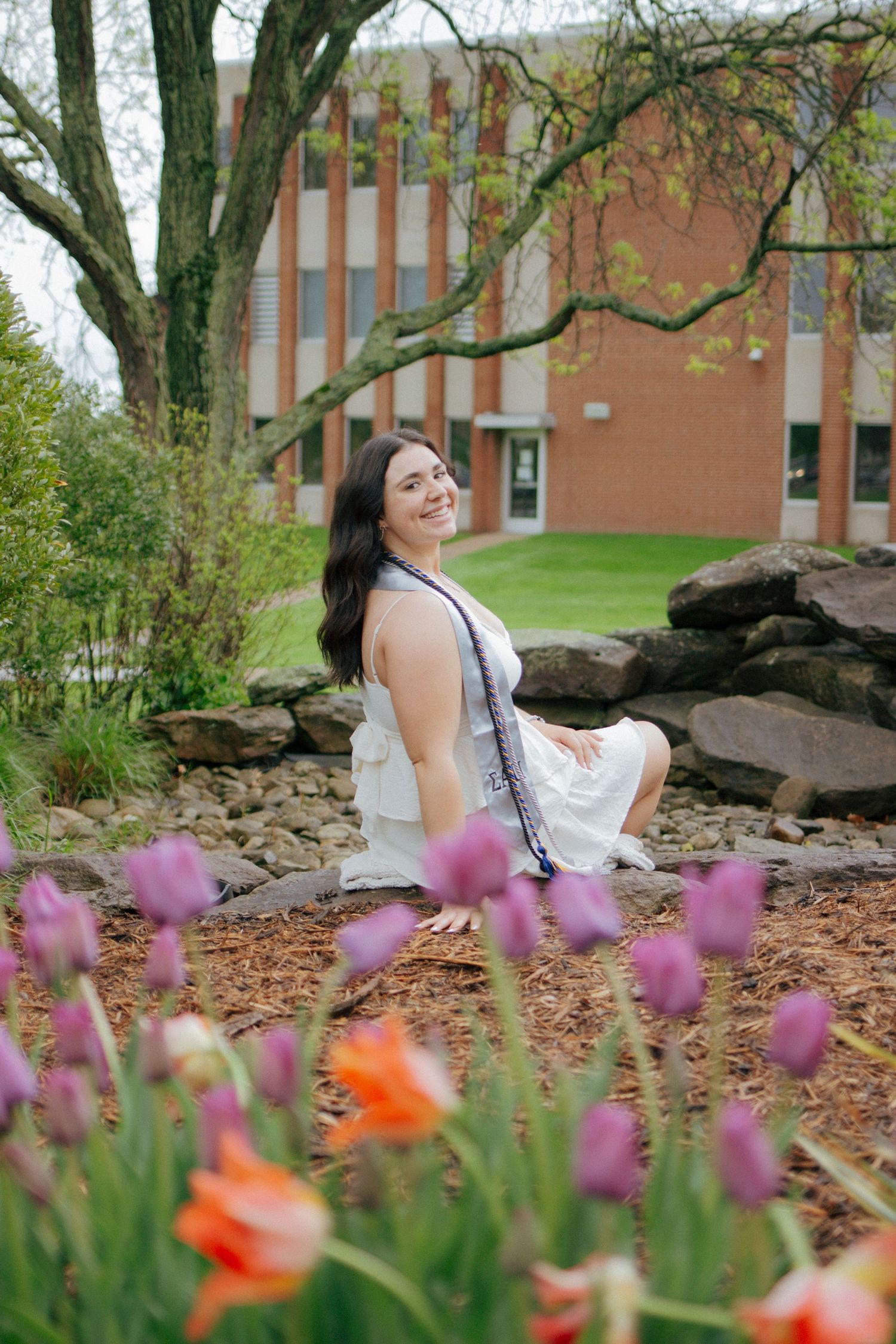 Smiling young woman in white dress poses among colorful tulips in a garden near a brick building.
