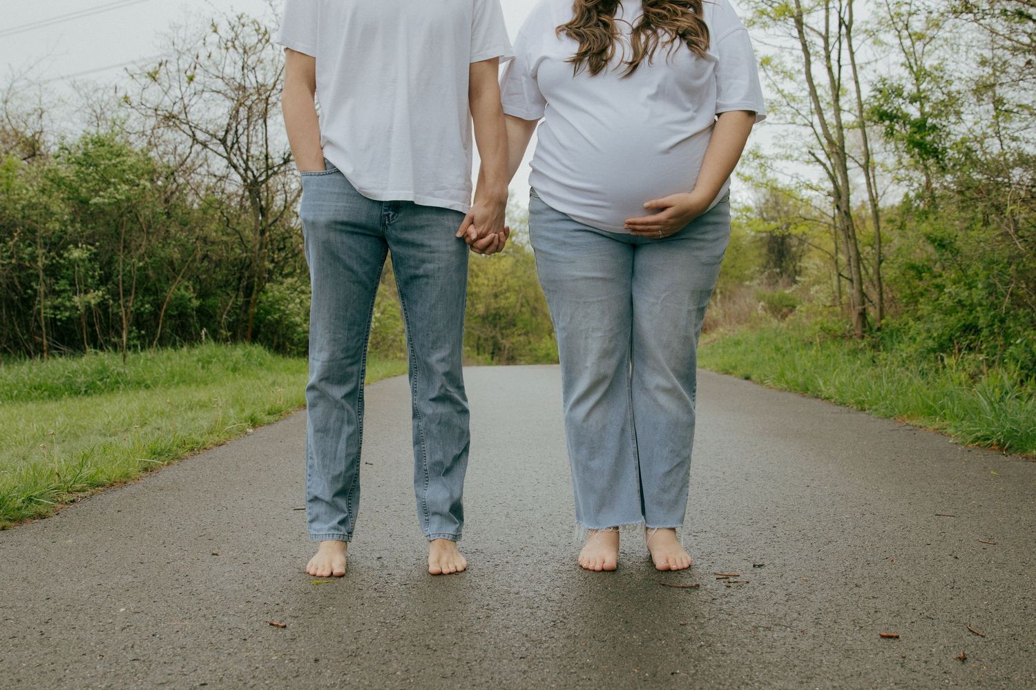 Barefoot pregnant couple holding hands standing on a paved road in Pittsburgh at Settlers Cabin Park