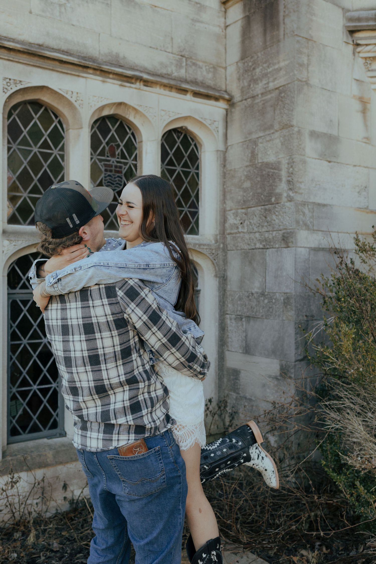 Man in plaid shirt lifts laughing woman in denim jacket outside historic stone building.