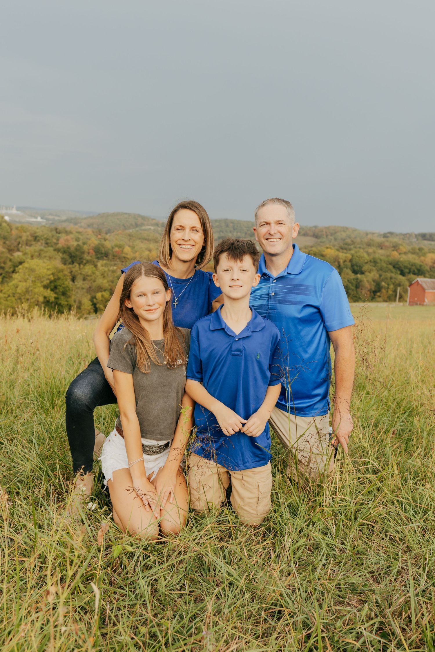 Family of four poses together in a green field with rolling hills behind them.