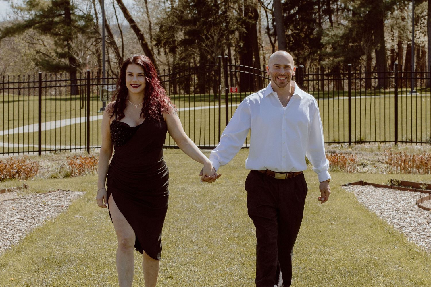 Couple holding hands walking through a garden path with green lawn and iron fence.