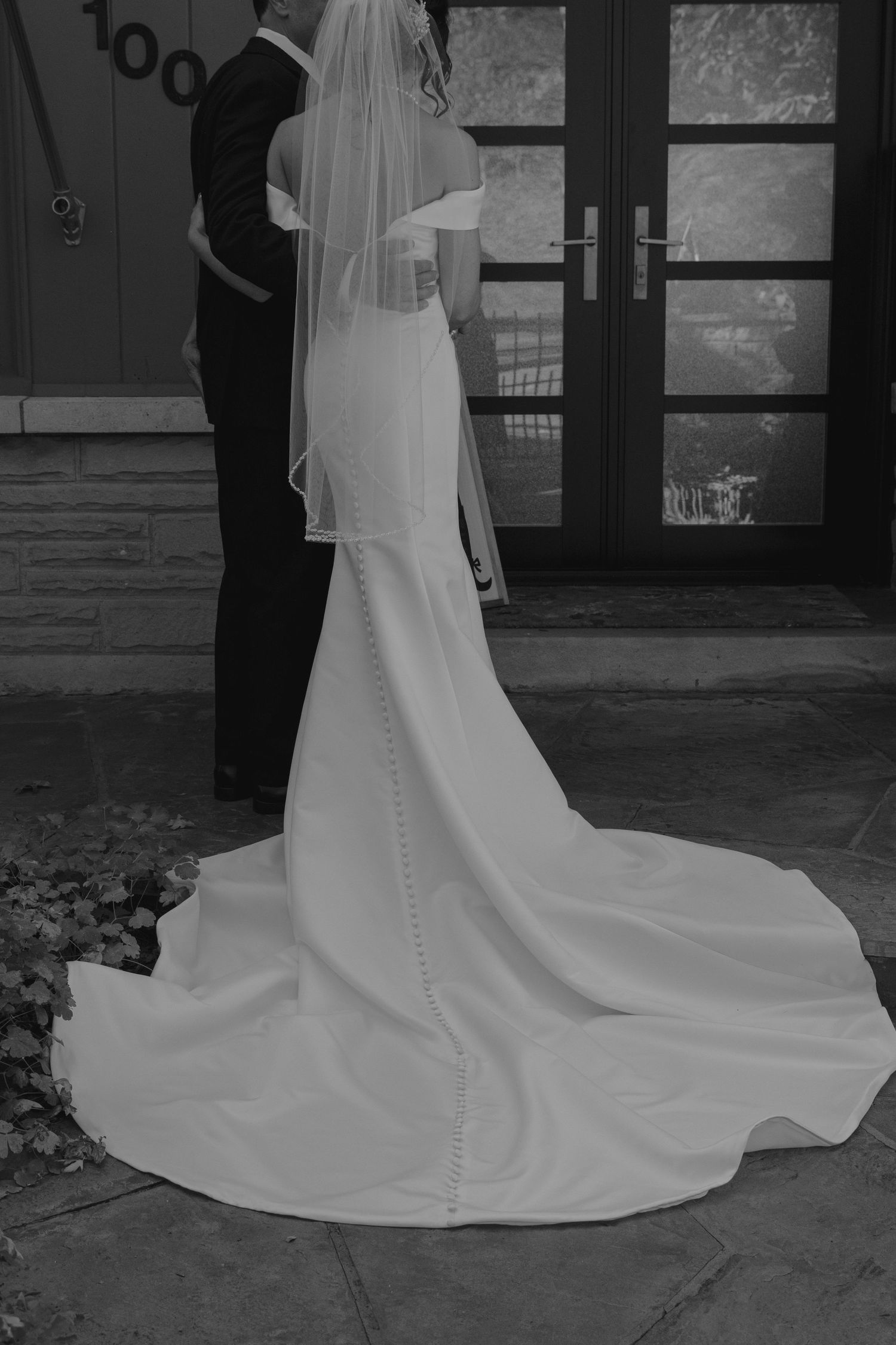 Black and white photo of bride in fitted gown with long train and veil standing at venue entrance. Pittsburgh wedding photography.