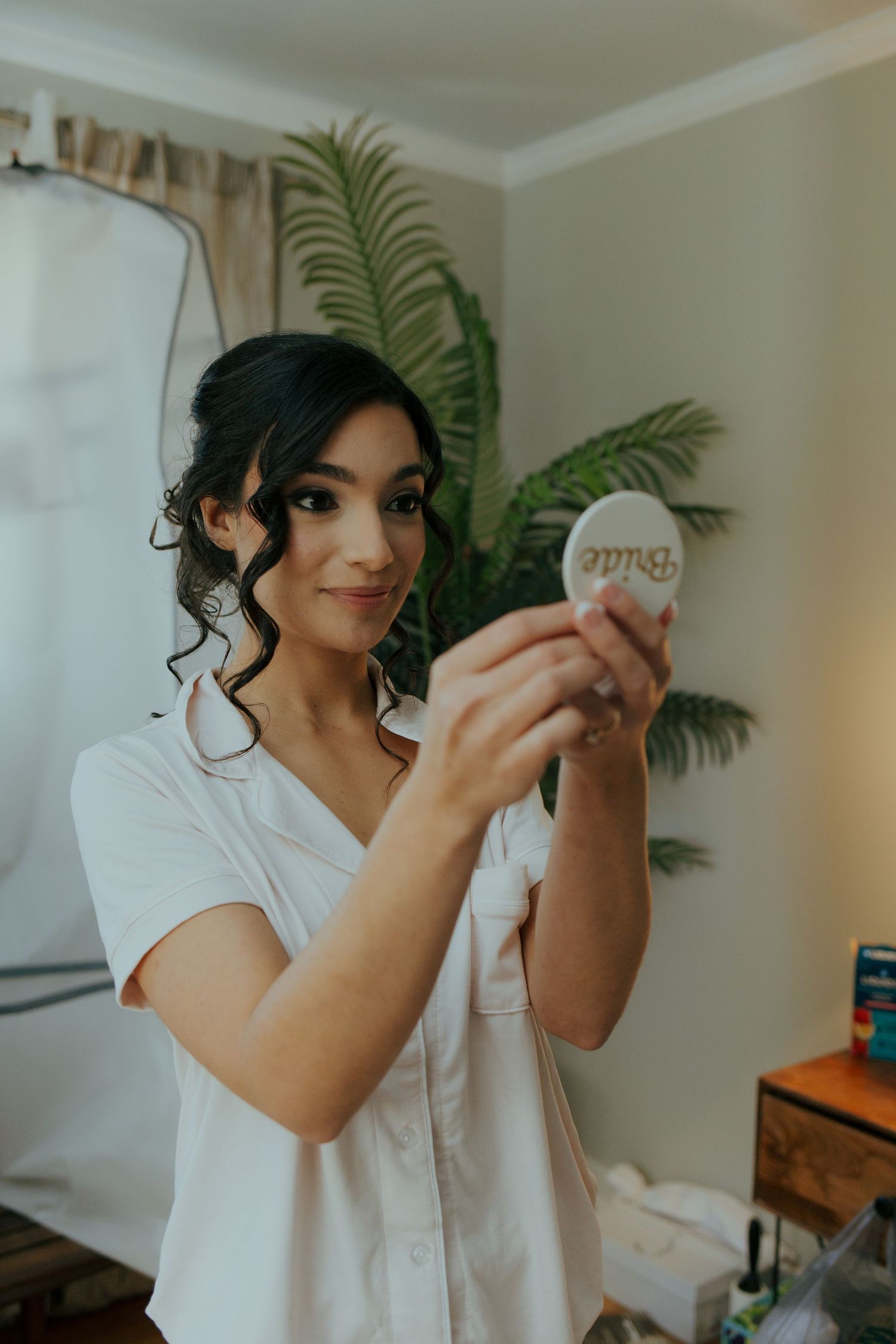 Beautiful dark-haired bride in white robe holding a decorative Bride mirror while getting ready on her wedding day.