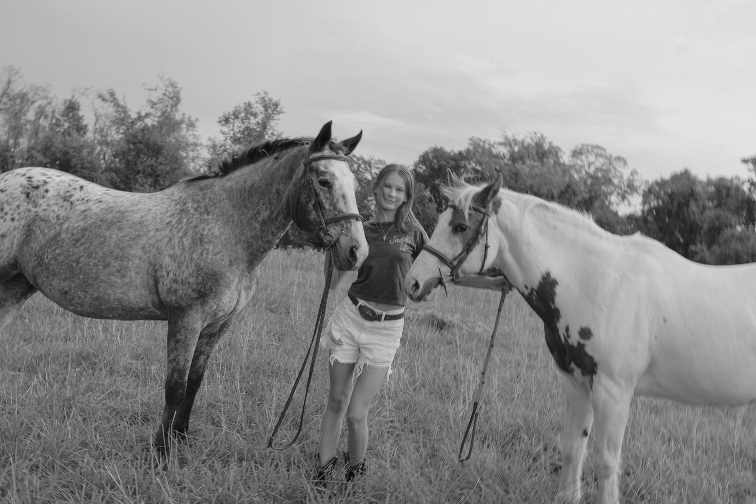 Young woman posing with two horses in a black and white outdoor field photo.