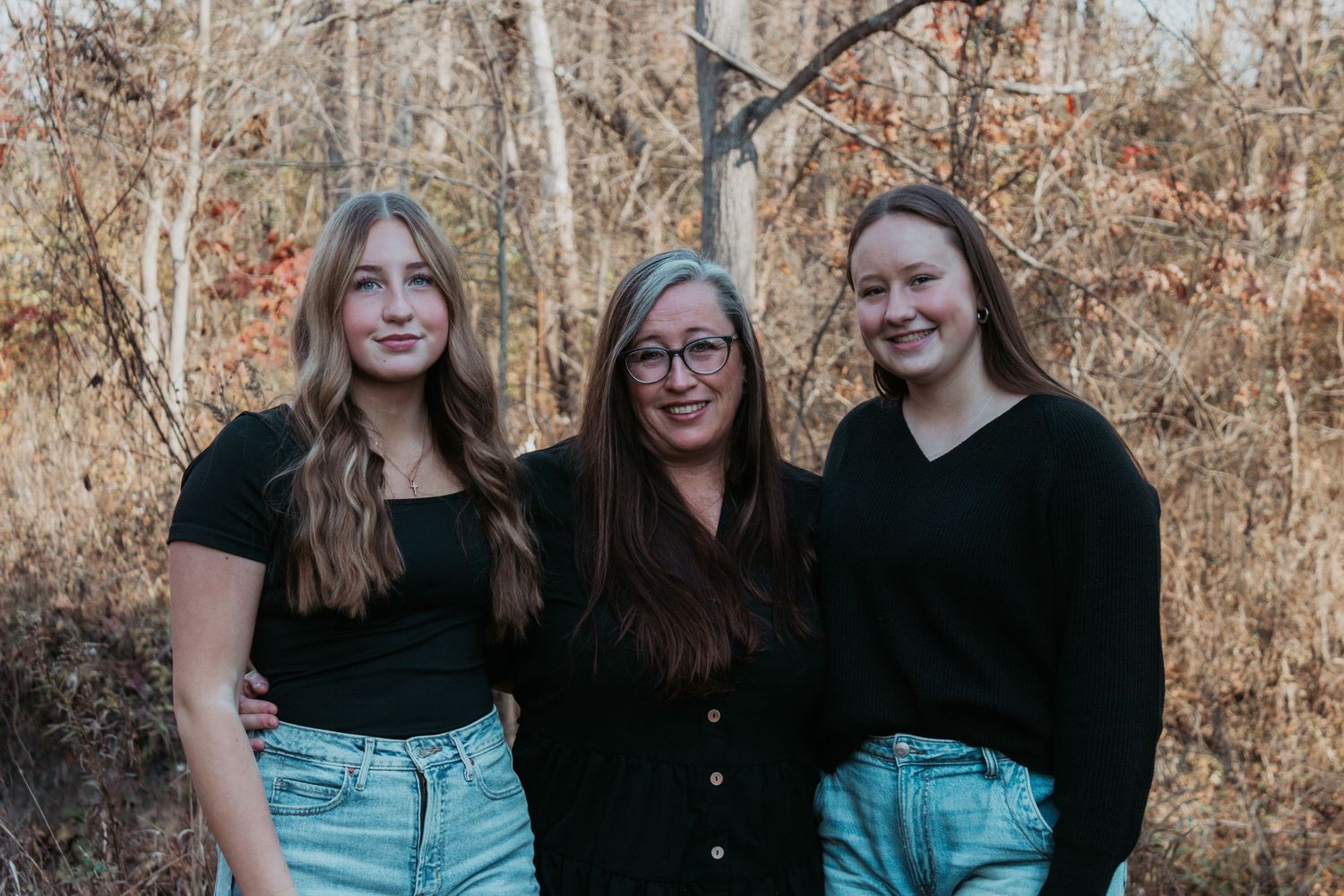 Three women in black outfits posing together outdoors among autumn foliage.