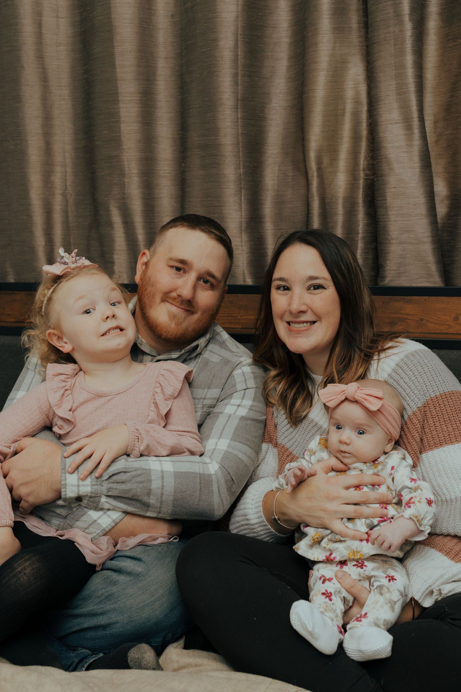 Happy family portrait with parents holding two young daughters in pink outfits on a cozy bed.
