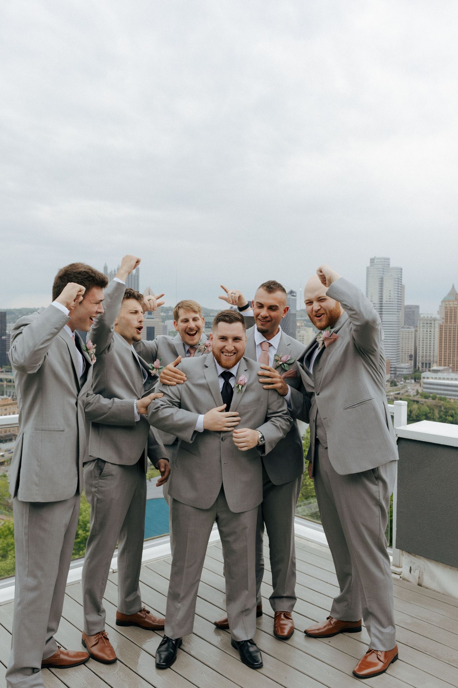 Groom and groomsmen in gray suits celebrate on rooftop with city skyline backdrop.