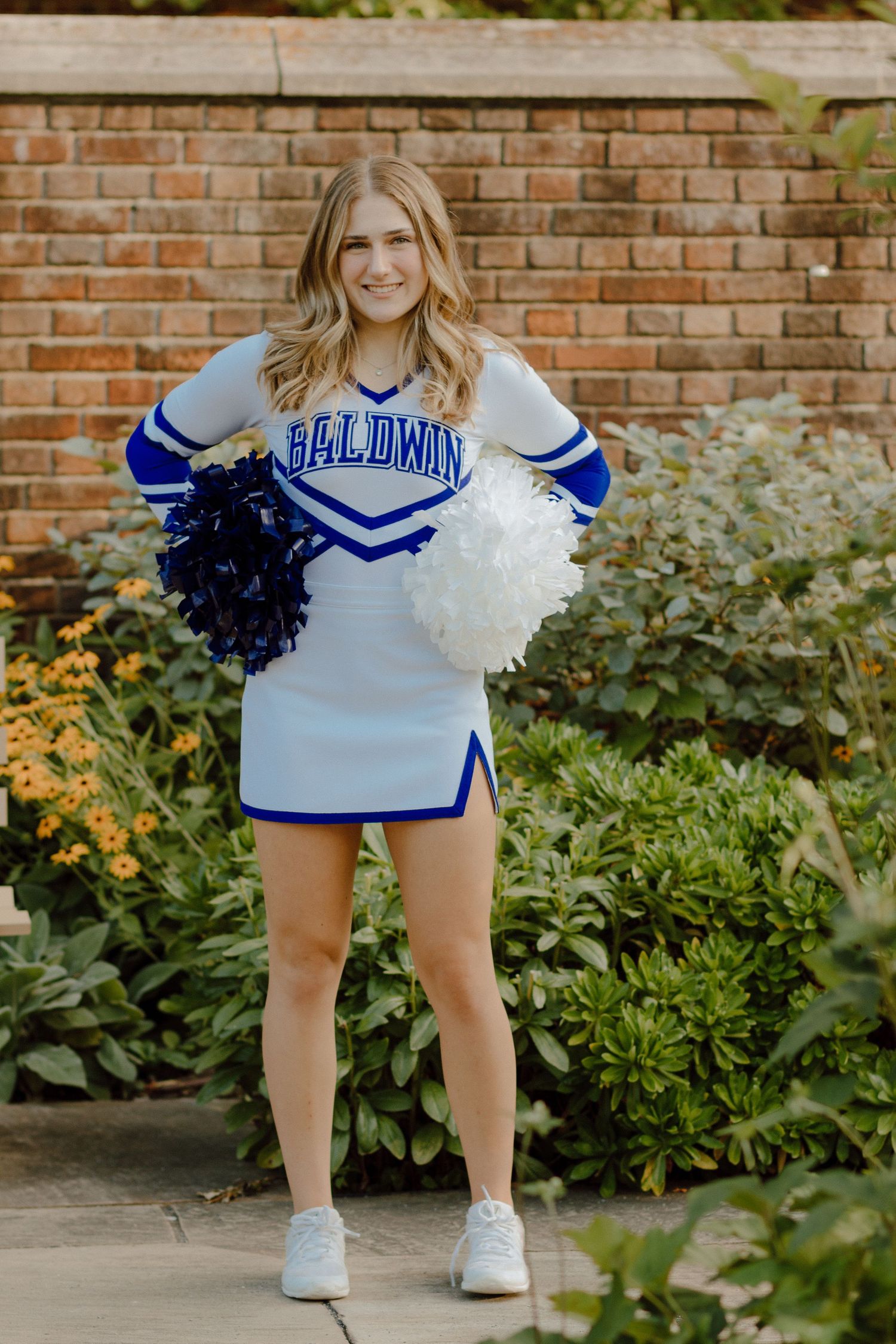 Cheerleader in Baldwin blue and white uniform poses with pom poms in front of brick wall and greenery.