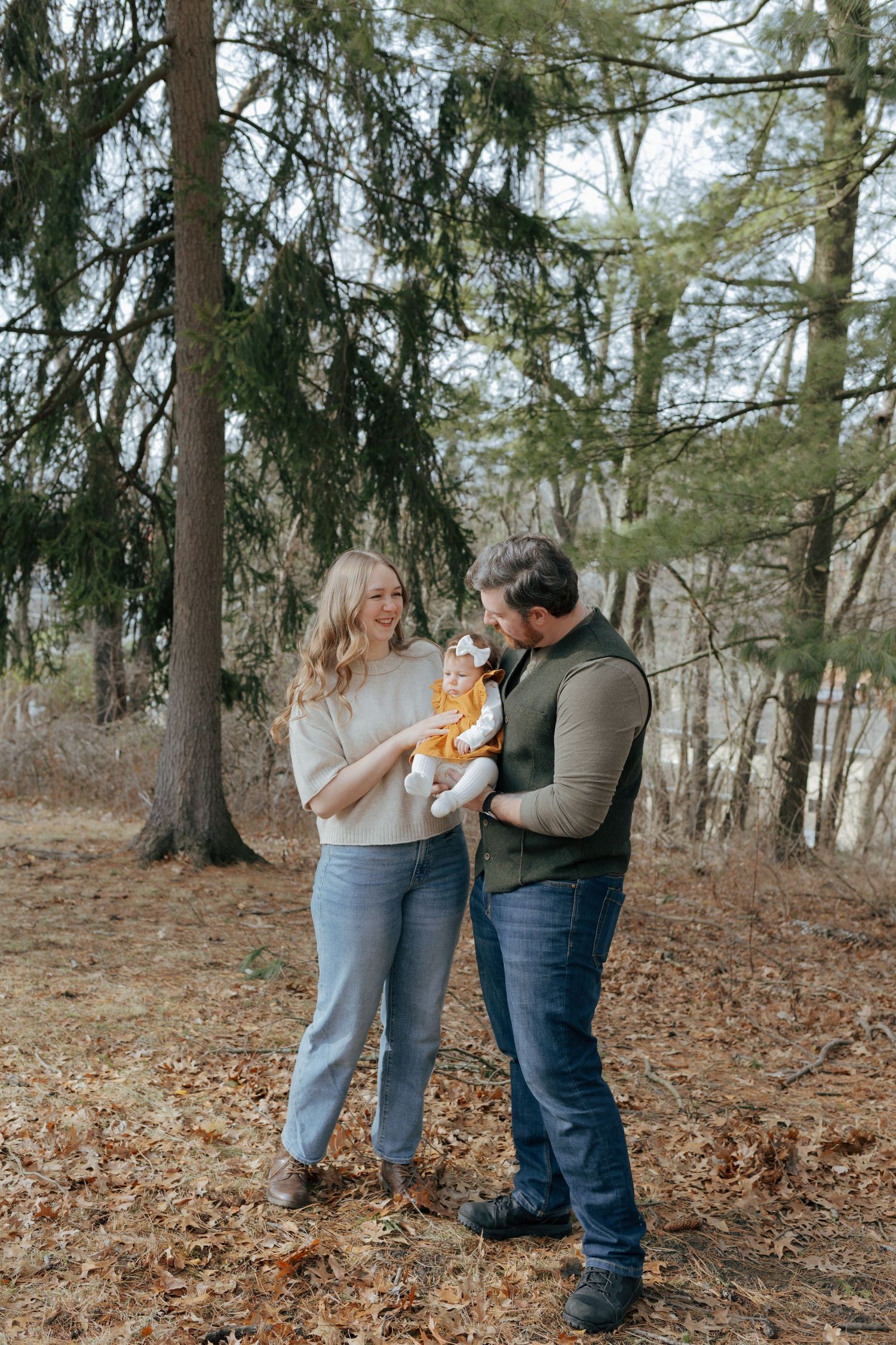 A young couple smiles while holding their baby outdoors in a forest setting with fallen leaves on the ground.