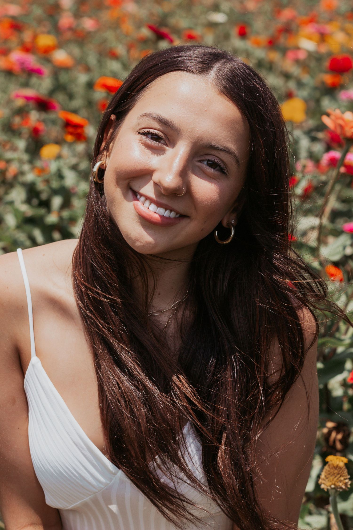 Smiling young woman with long dark hair in a white dress among colorful flowers.