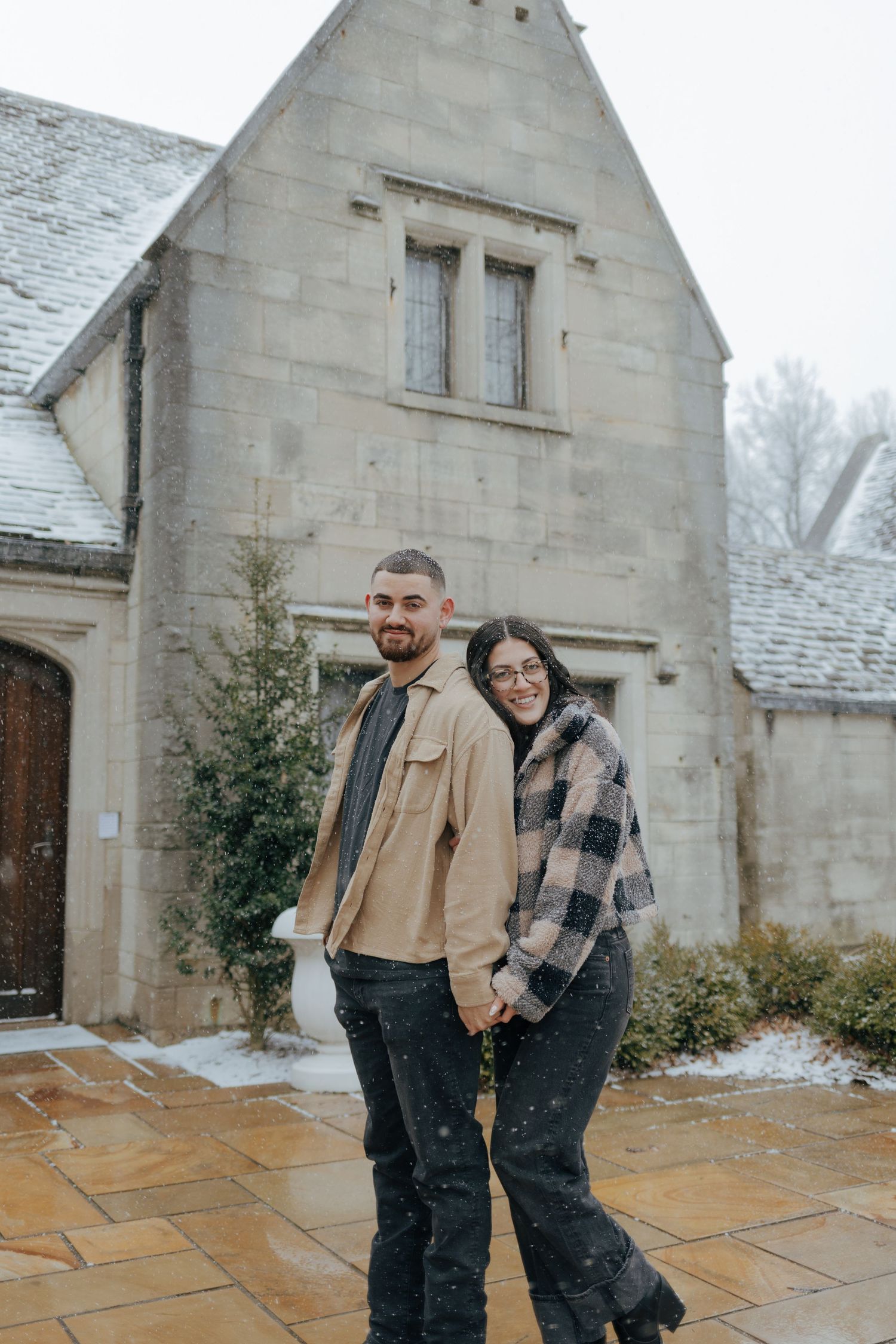 Smiling couple holding hands in front of a snow-dusted stone manor house in winter.