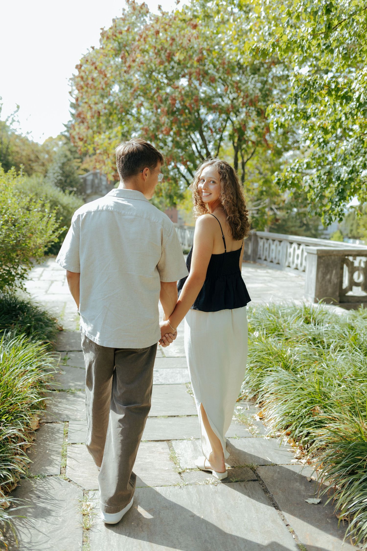Engaged couple walking hand in hand on a sunny garden path surrounded by lush greenery.