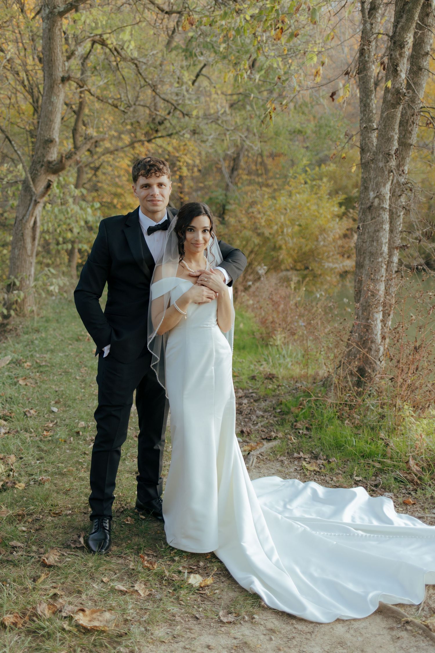 Elegant bride and groom posing together in an autumn forest, wearing white gown with long train and black tuxedo.