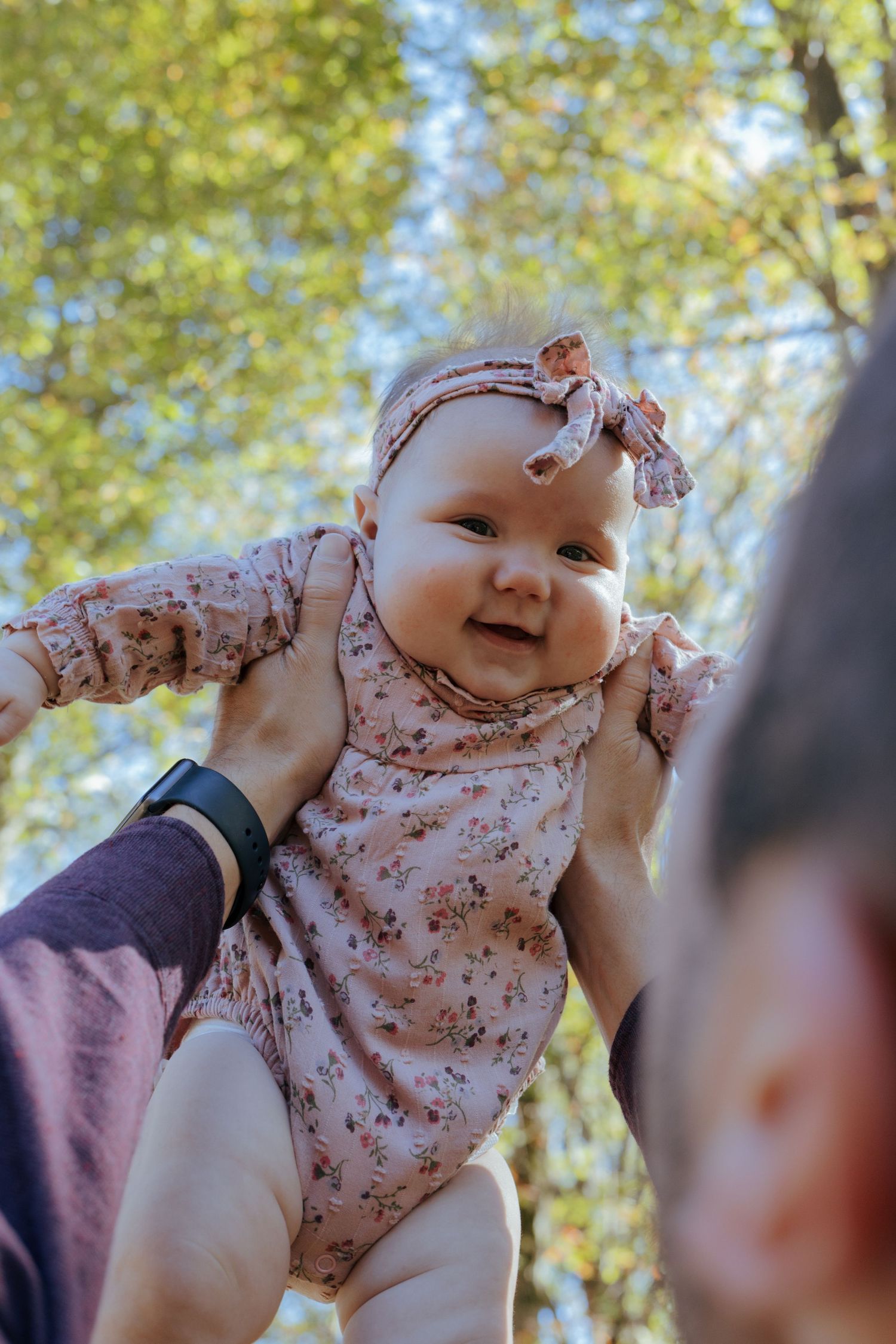 Smiling baby girl in floral onesie and bow headband being lifted up outdoors among green trees. Pittsburgh newborn session.