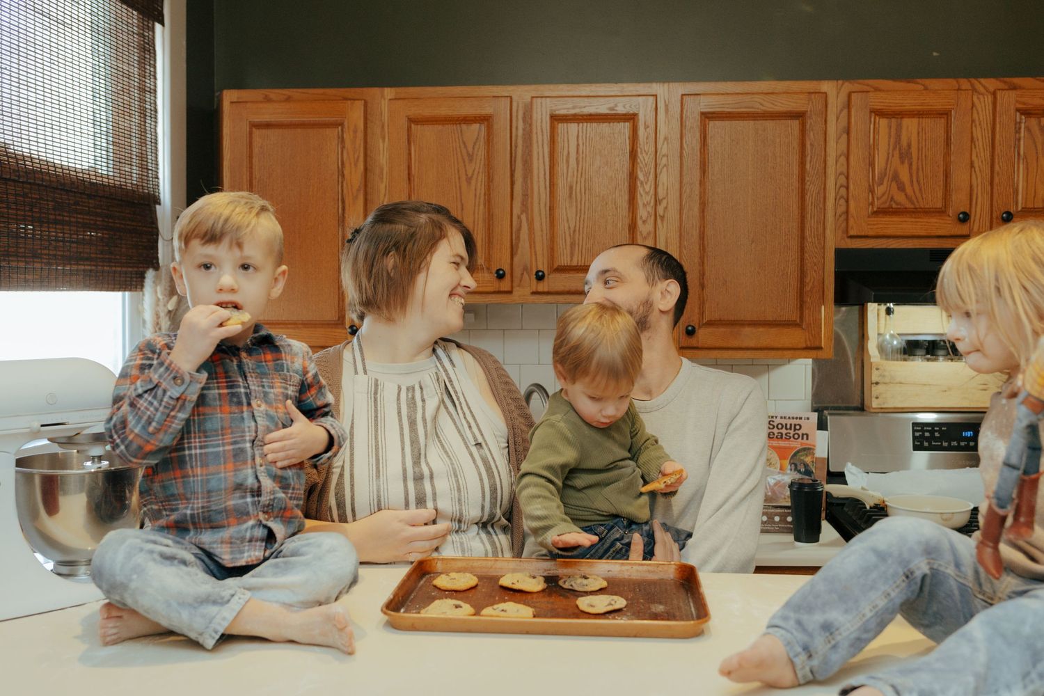 Happy family sitting on kitchen counter enjoying freshly baked cookies on a baking sheet.
