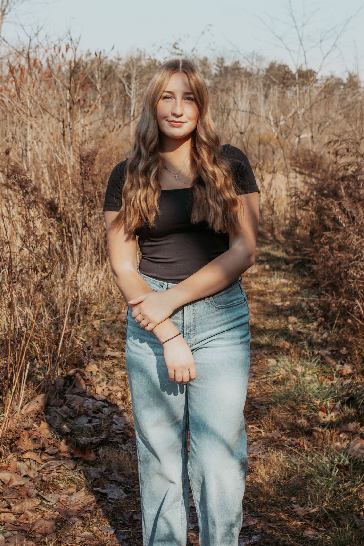 Teen girl with long wavy hair in black top and jeans posing in autumn landscape.