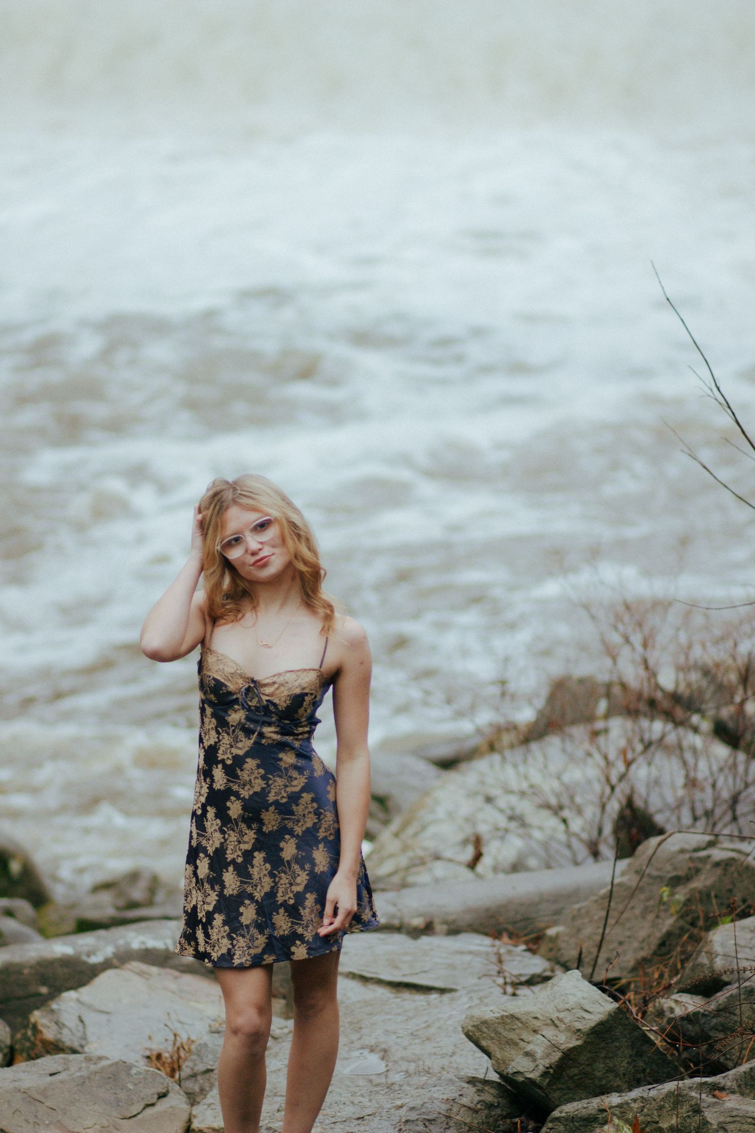 Young blonde woman in floral dress standing on rocks near rushing river waters.