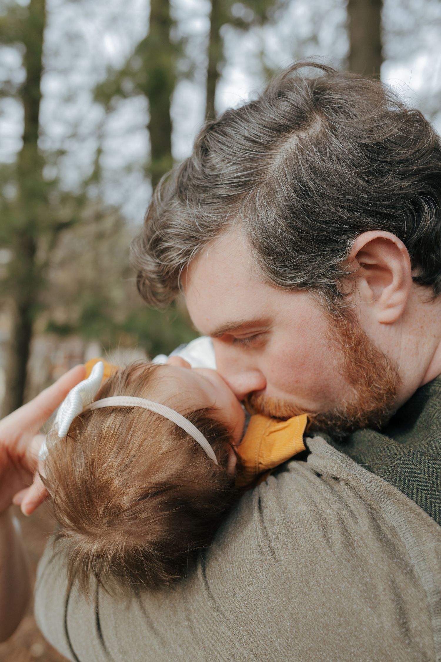 Loving father kissing baby daughter in mustard dress in a peaceful wooded outdoor setting.