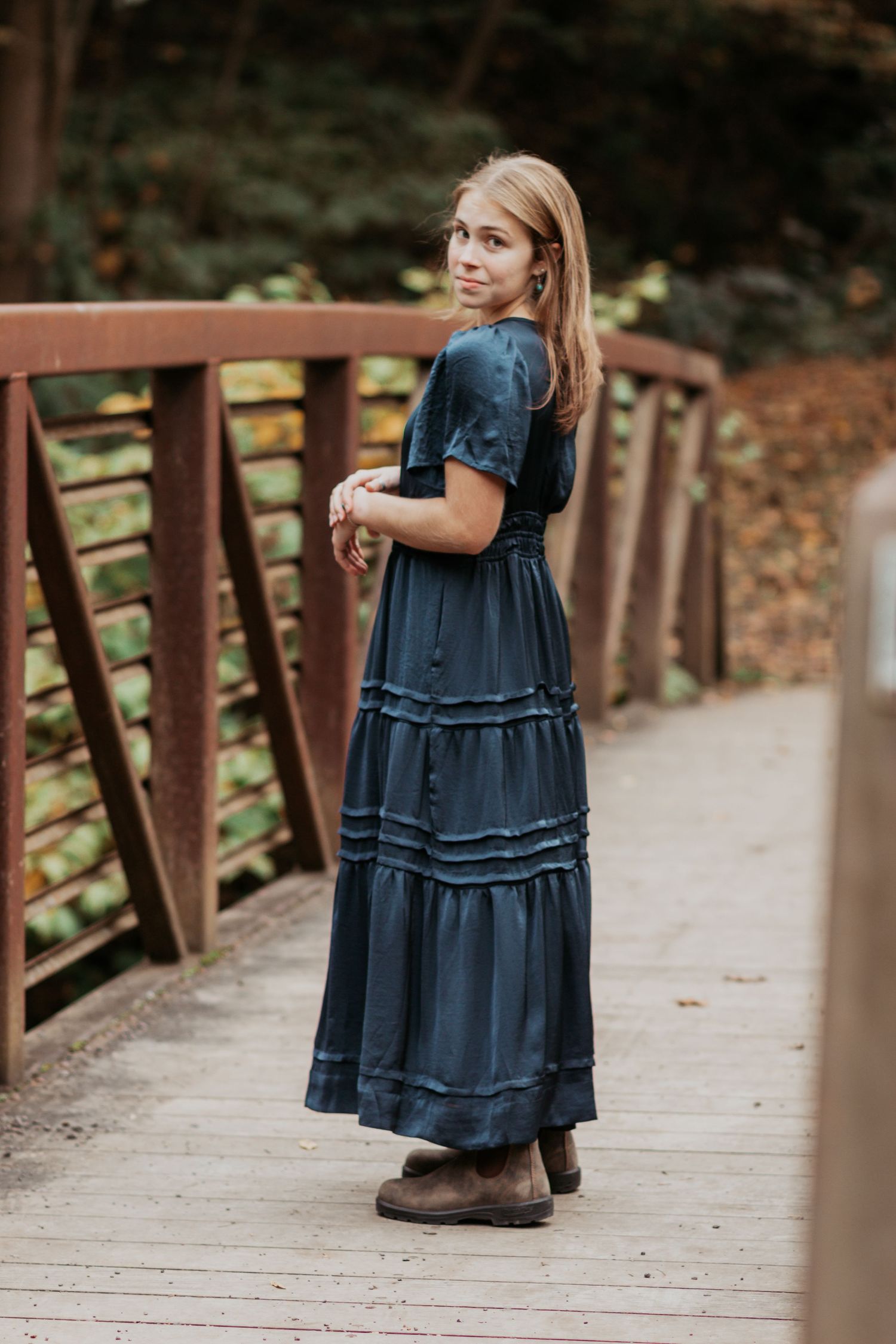 Young woman in navy blue maxi dress stands on wooden bridge surrounded by fall foliage.