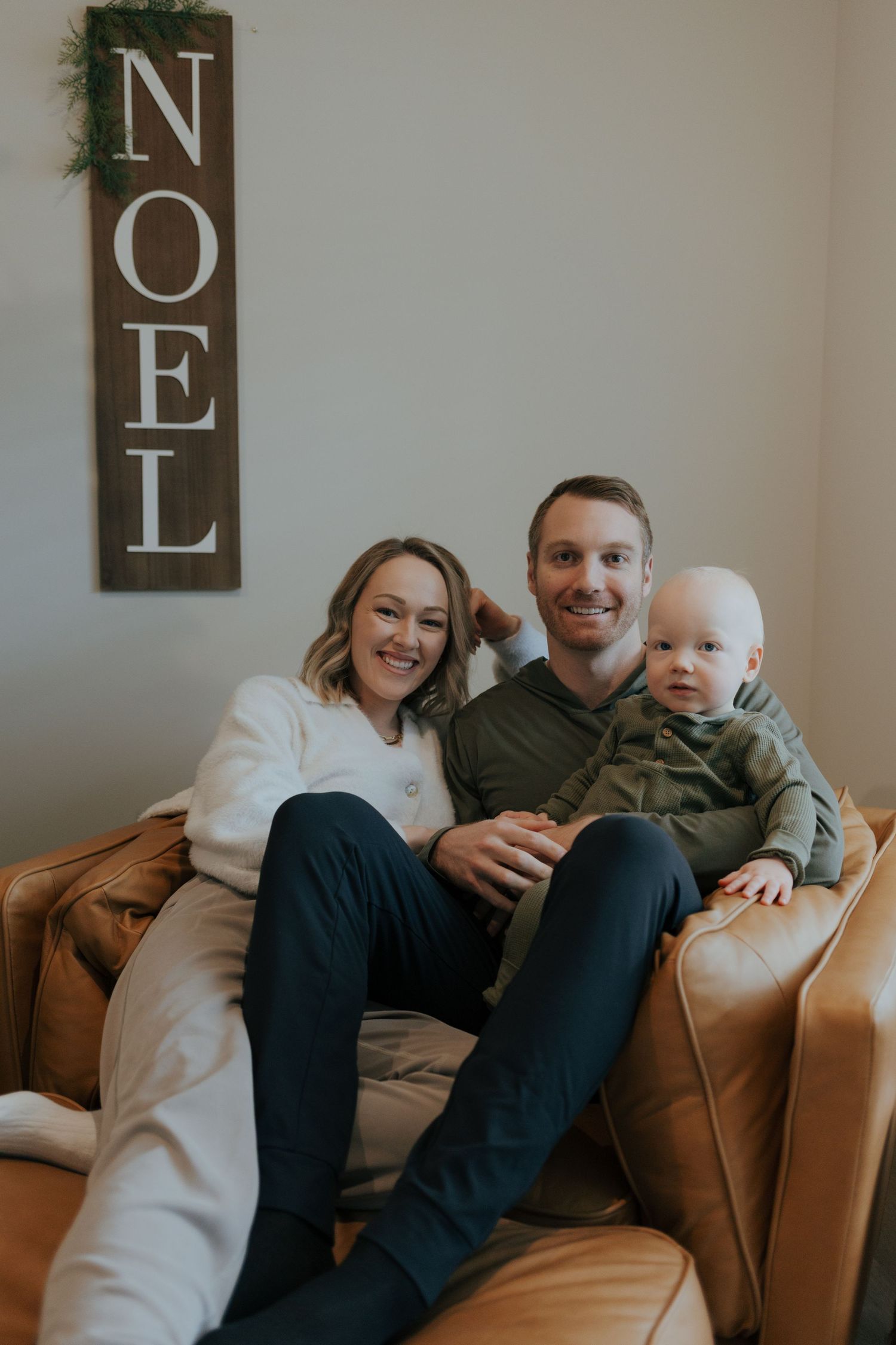 Happy young family with baby boy poses on leather couch beside a wooden NOEL Christmas sign.