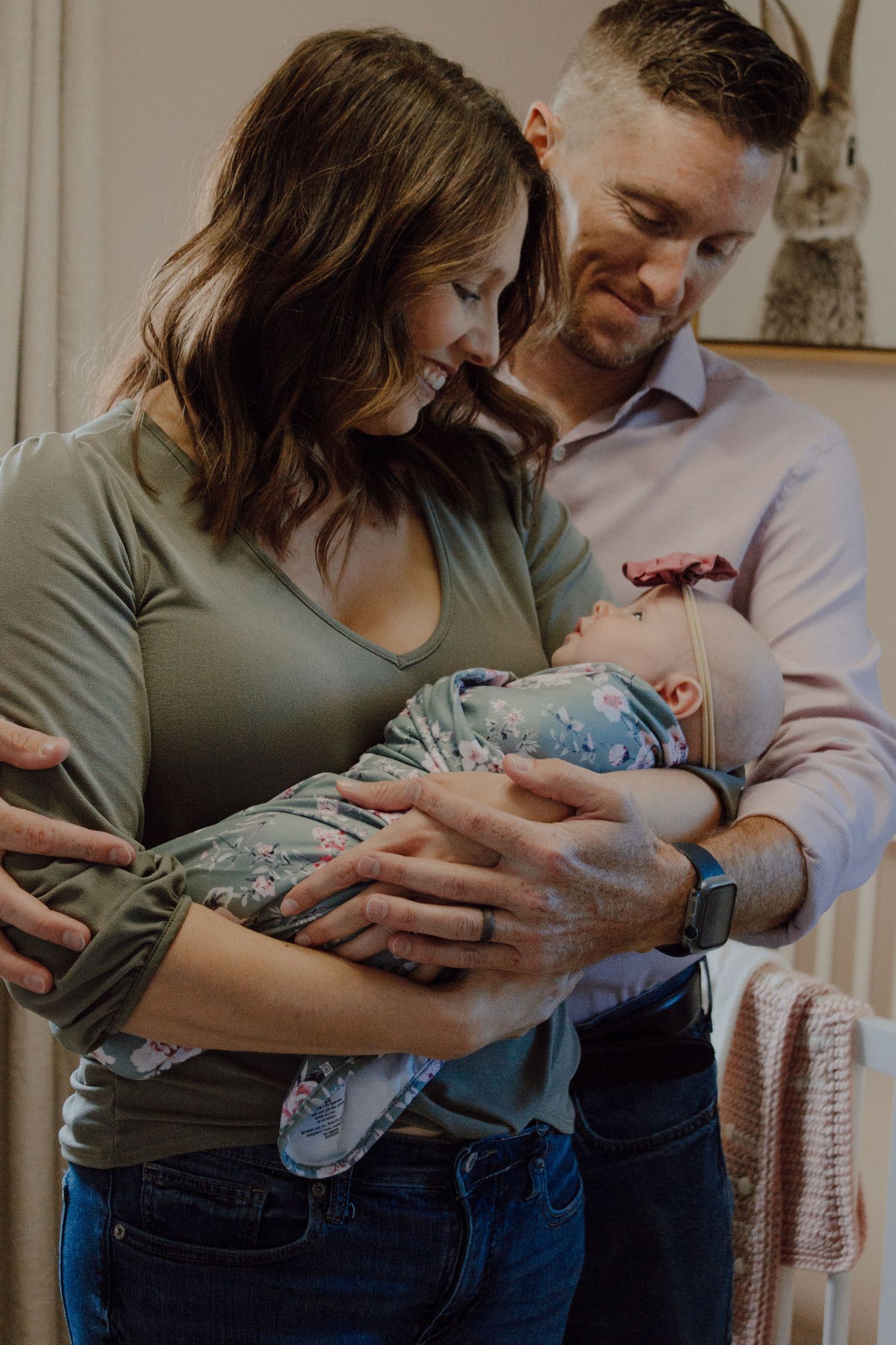 Loving parents smile down at their swaddled newborn baby in a warmly lit nursery room.