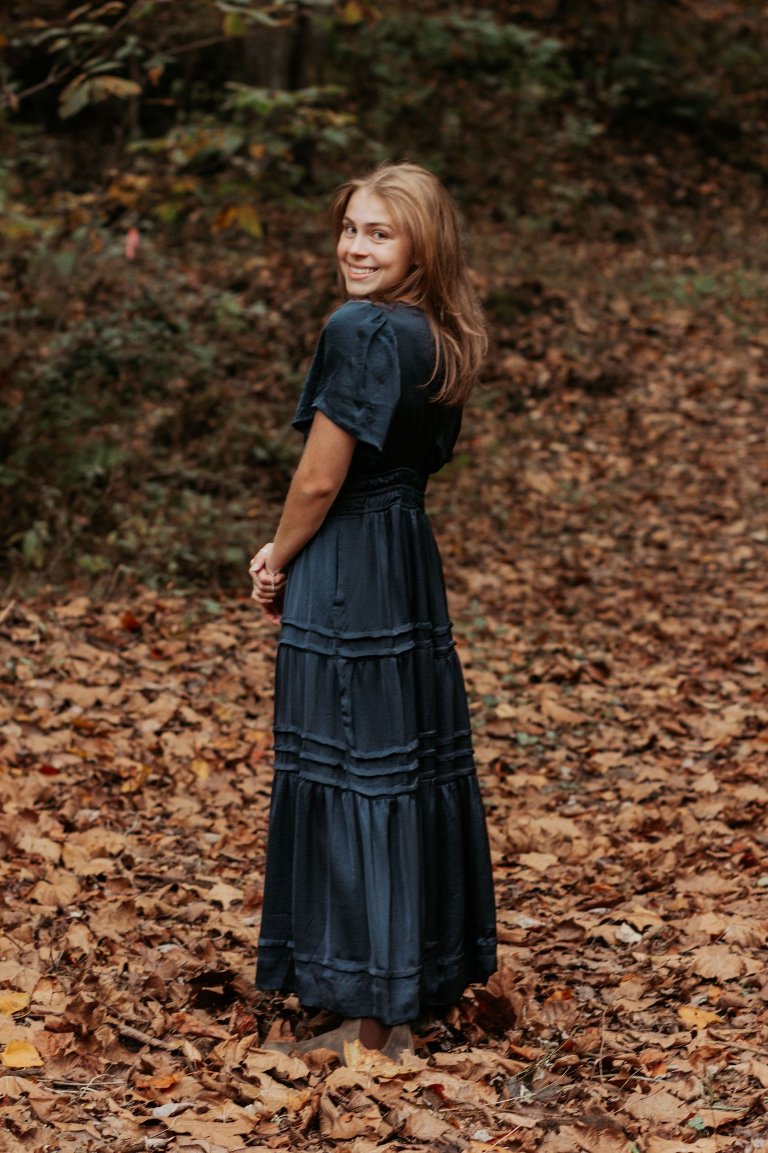 Young blonde woman in dark navy maxi dress standing among autumn fallen leaves.