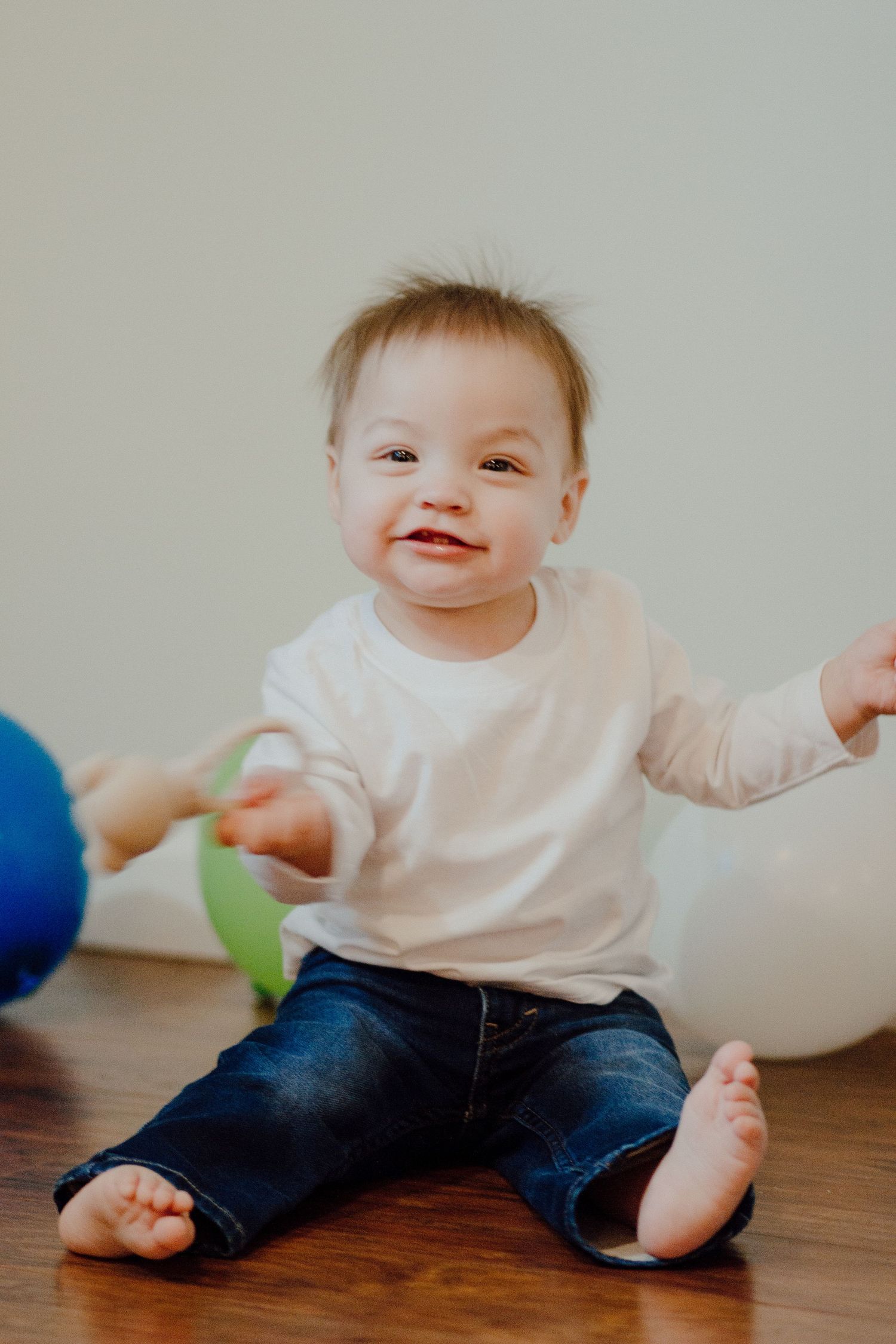 Smiling baby with Down syndrome sitting on floor reaching for green balloon at colorful birthday celebration.