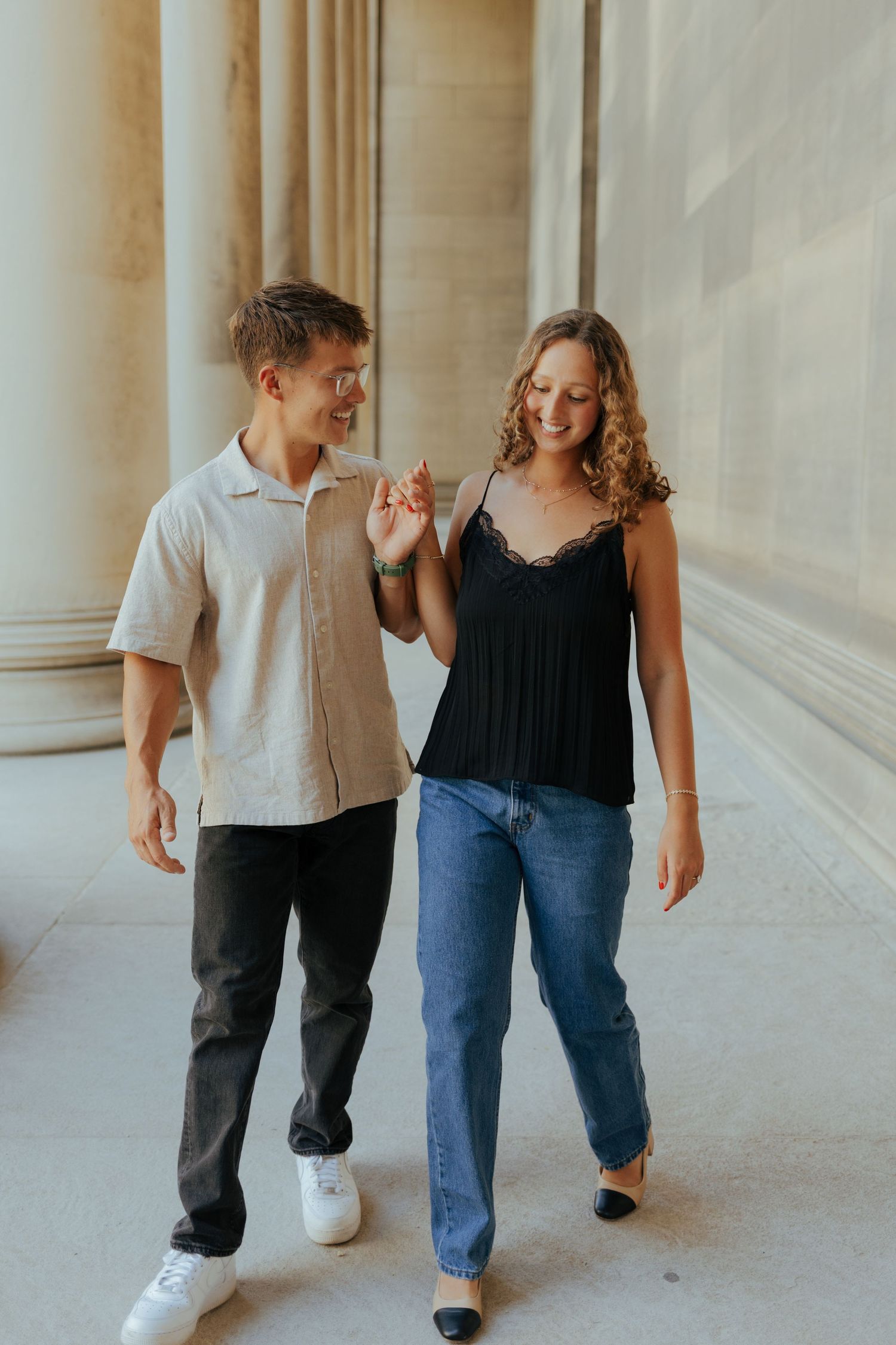 Happy engaged couple walking hand in hand between tall stone columns in a classic building.