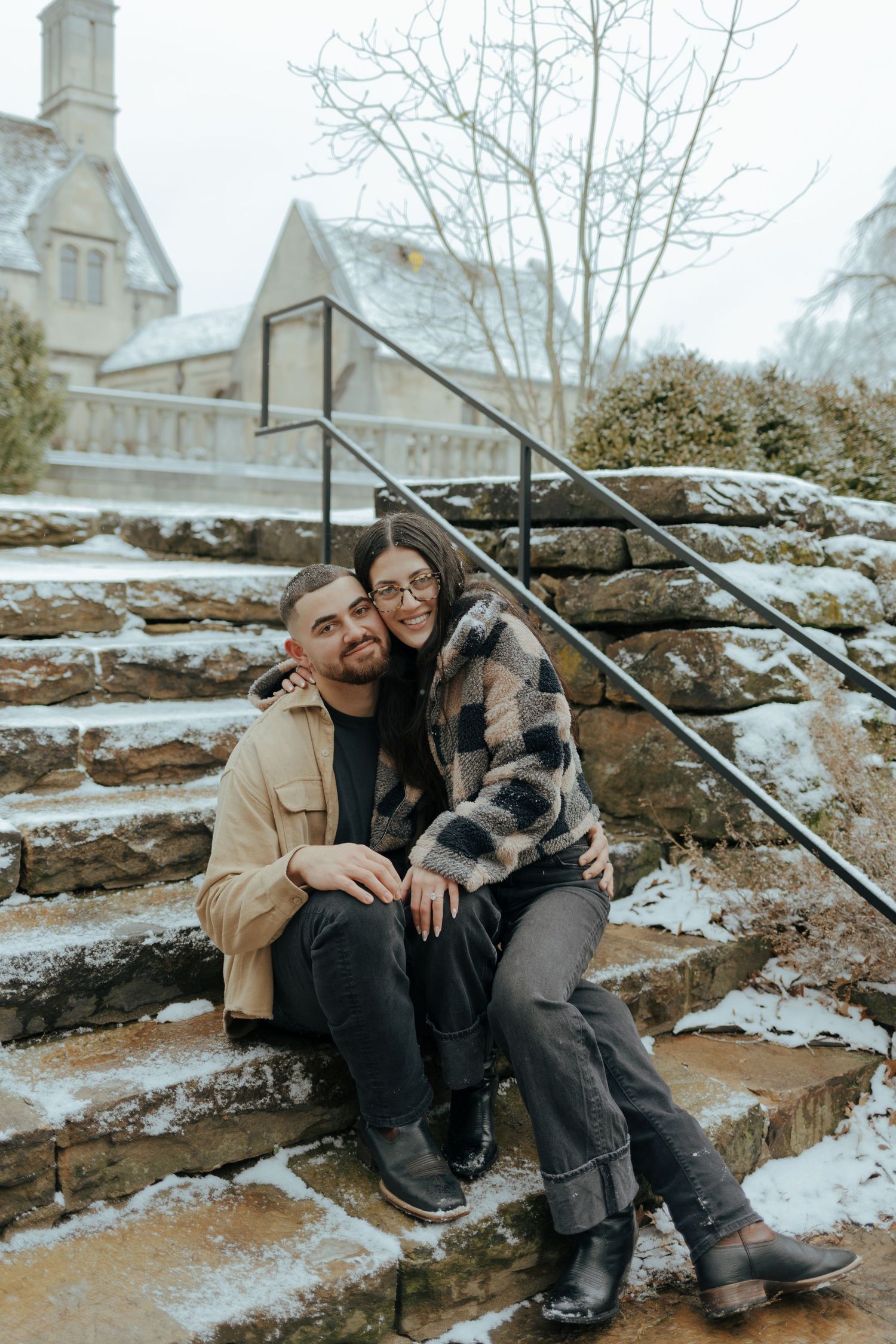 Couple sitting on snowy stone steps embracing in winter, wearing casual coats at Hartwood Acres in Pittsburgh.