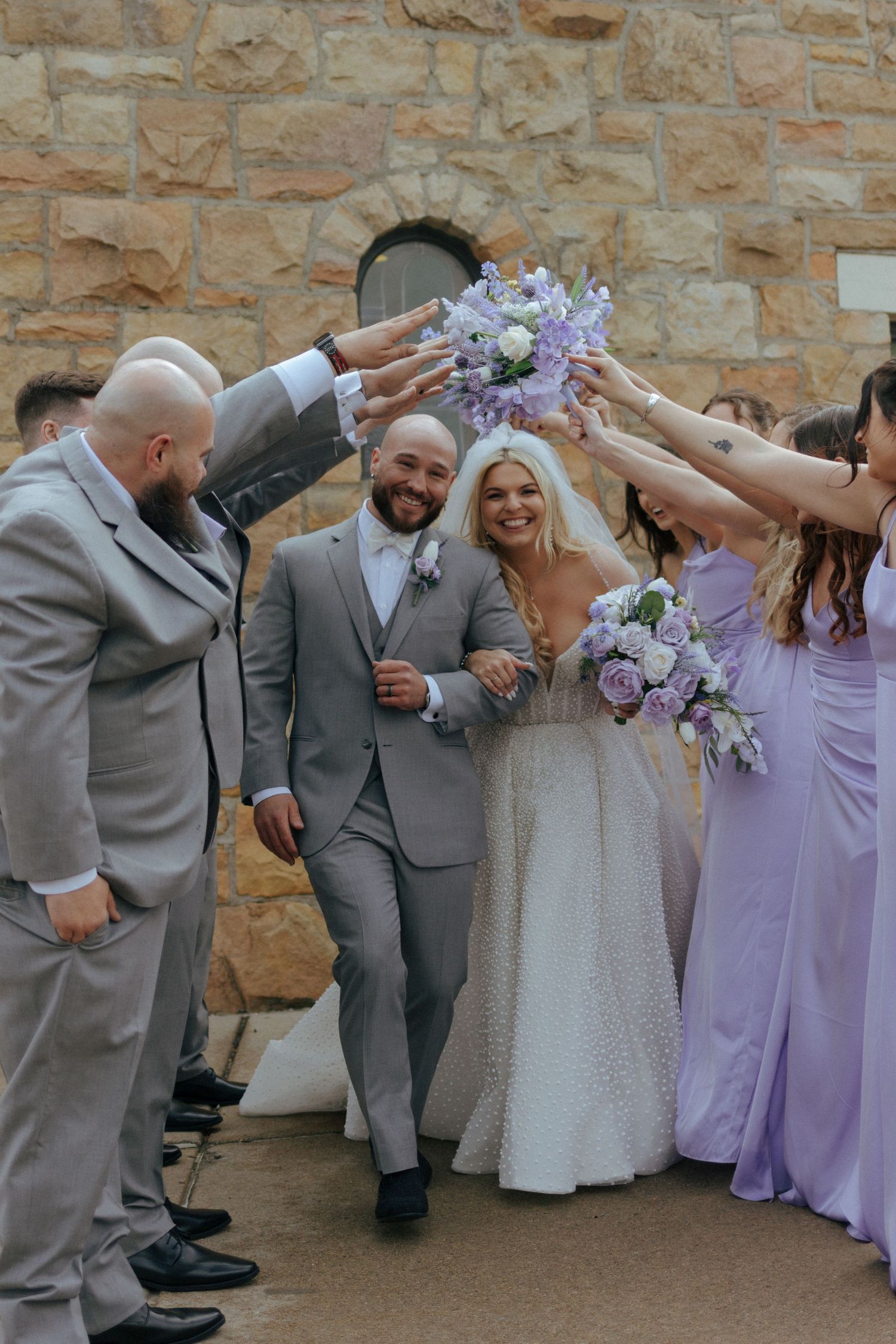 Bride and groom walking under arch of raised bouquets with wedding party in purple dresses in Pittsburgh