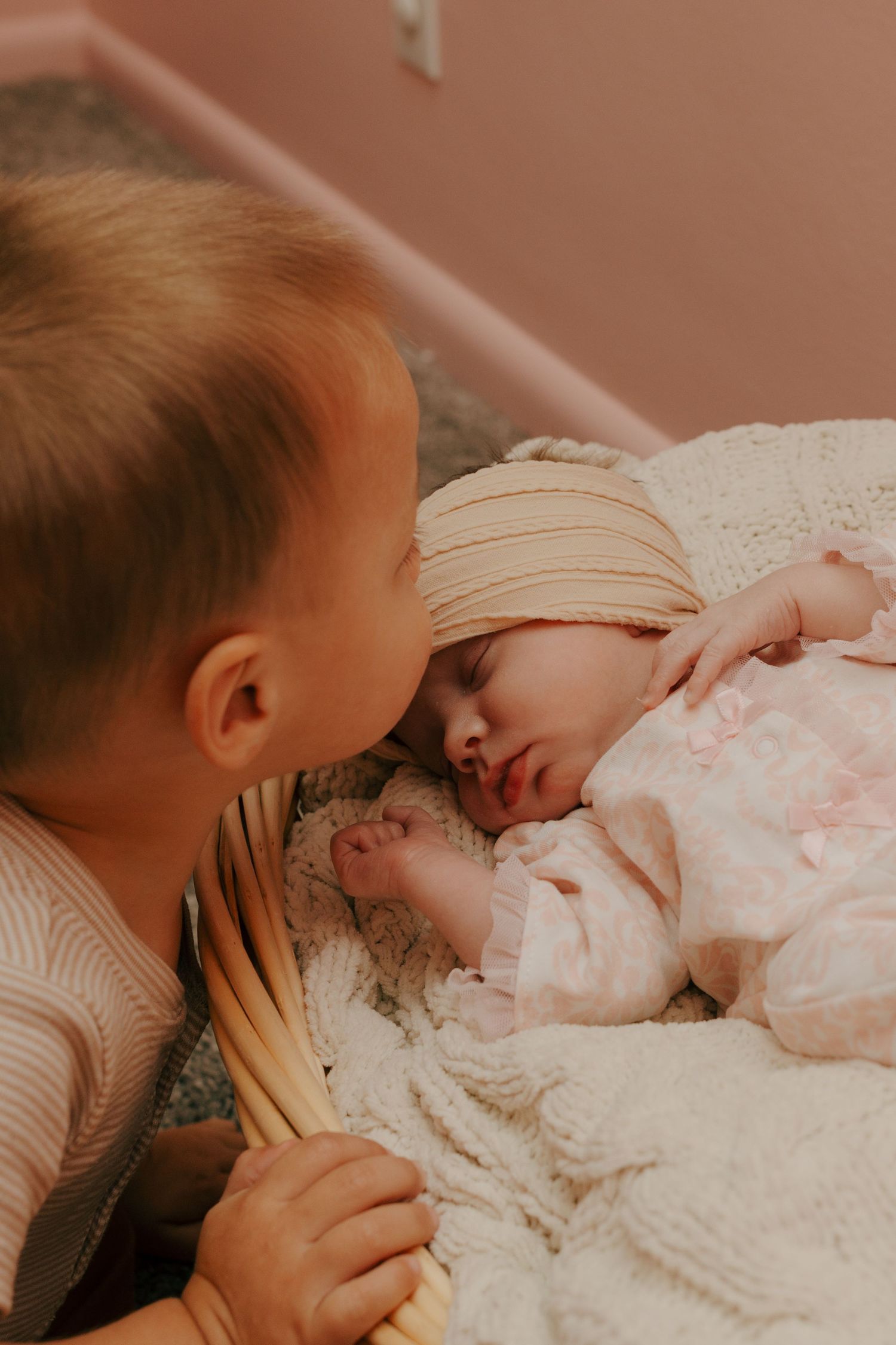 Young toddler gently leaning in to kiss sleeping newborn baby sister lying in wicker basket.