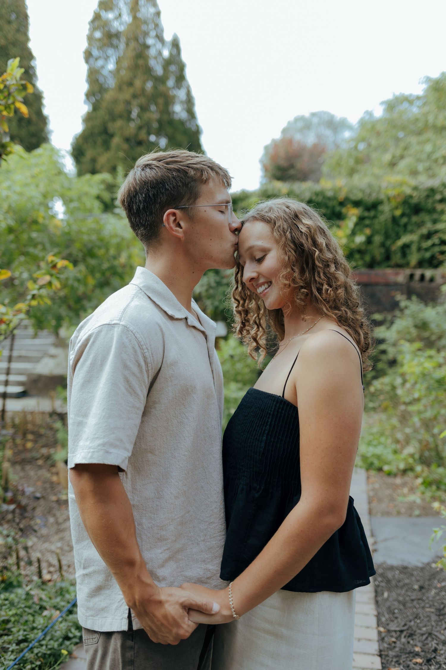 Young man kissing smiling woman on the forehead while holding hands in a lush garden.
