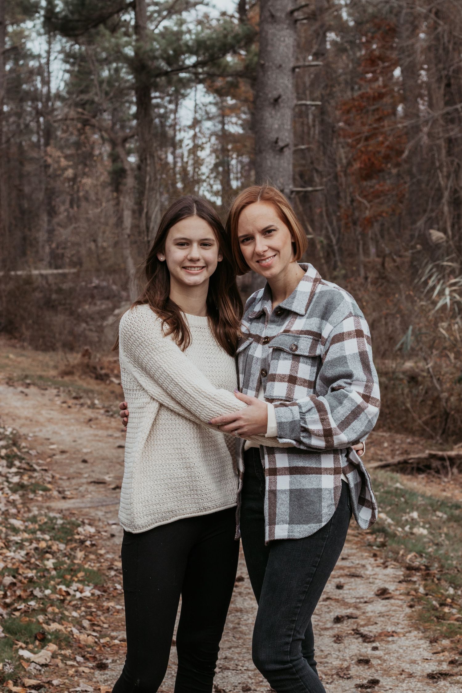 Mother and teenage daughter embracing on autumn forest path, smiling at camera.