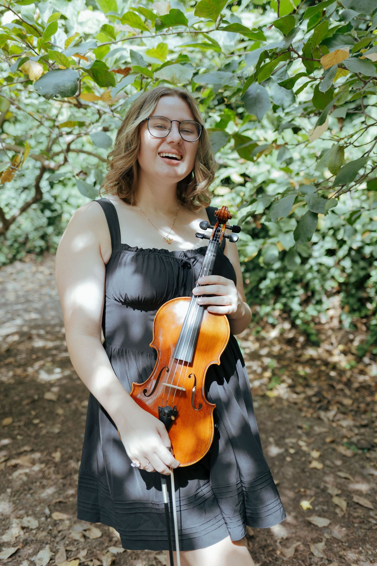 Smiling young woman in black dress holds violin while standing among lush green garden foliage.
