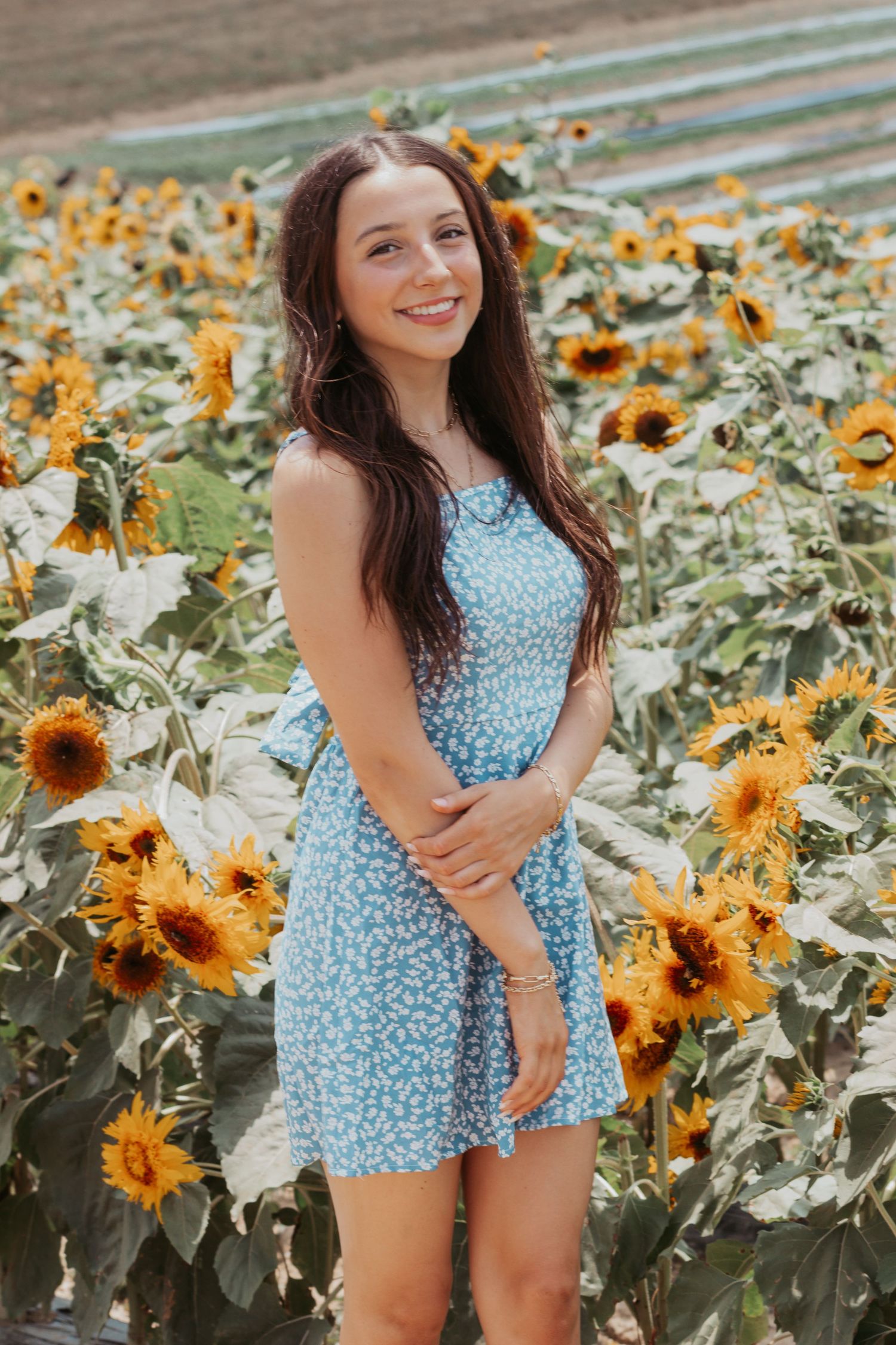 Smiling young woman in blue floral dress standing in a sunny sunflower field.
