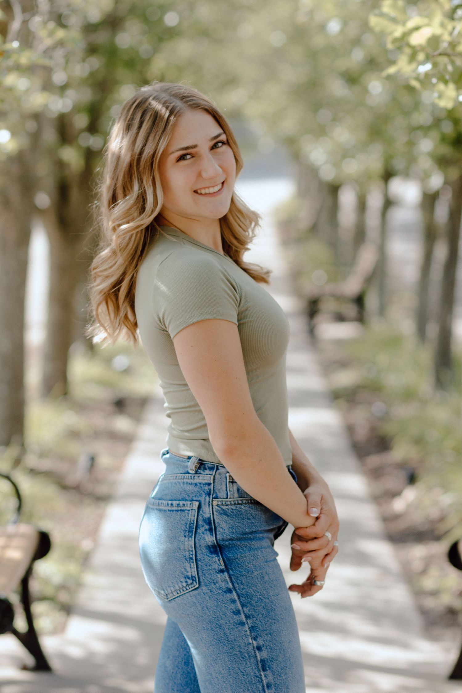 Smiling young woman in sage green top and jeans posing on a tree-lined path.