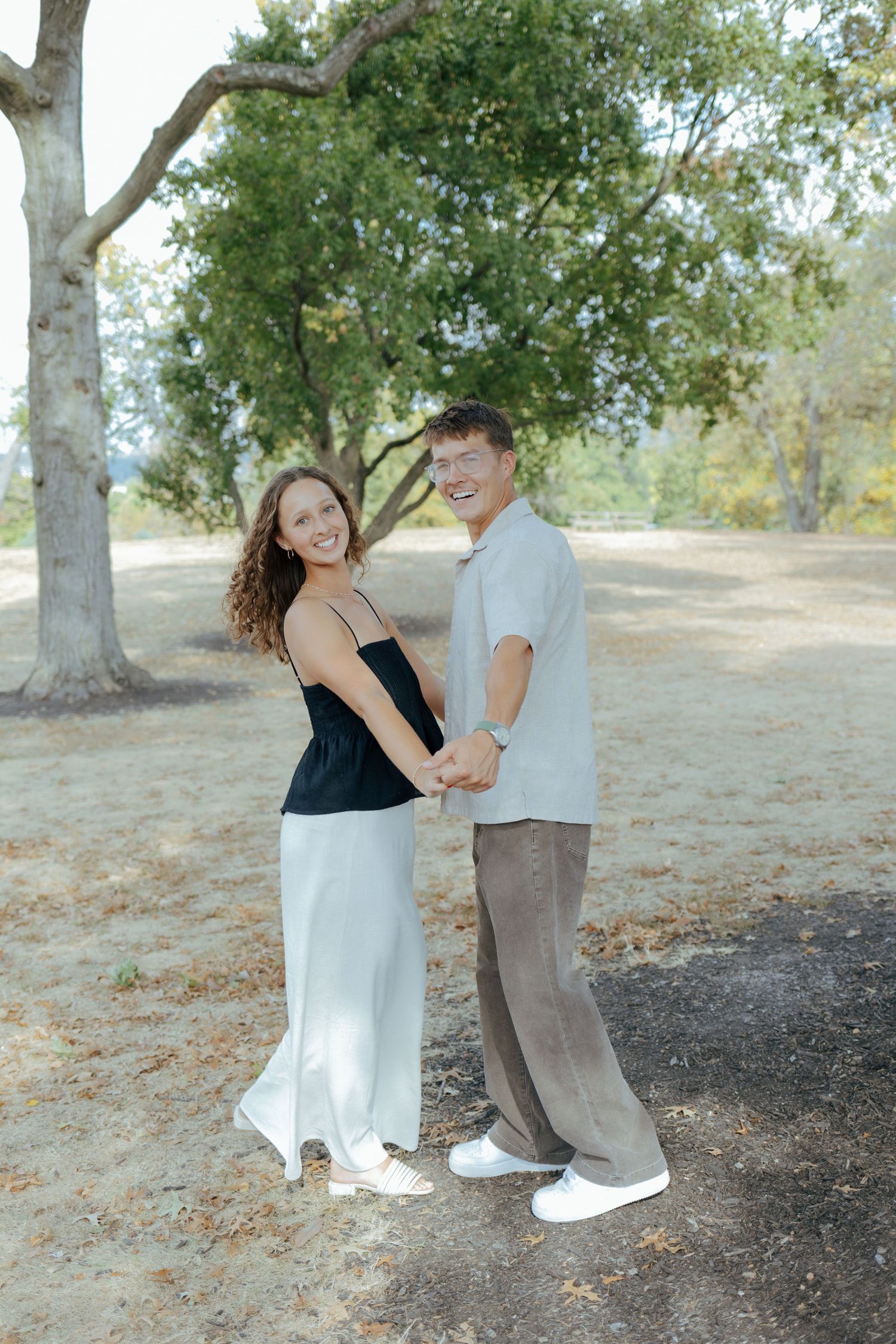 Happy couple holding hands and smiling under large trees in a sunlit outdoor park setting.