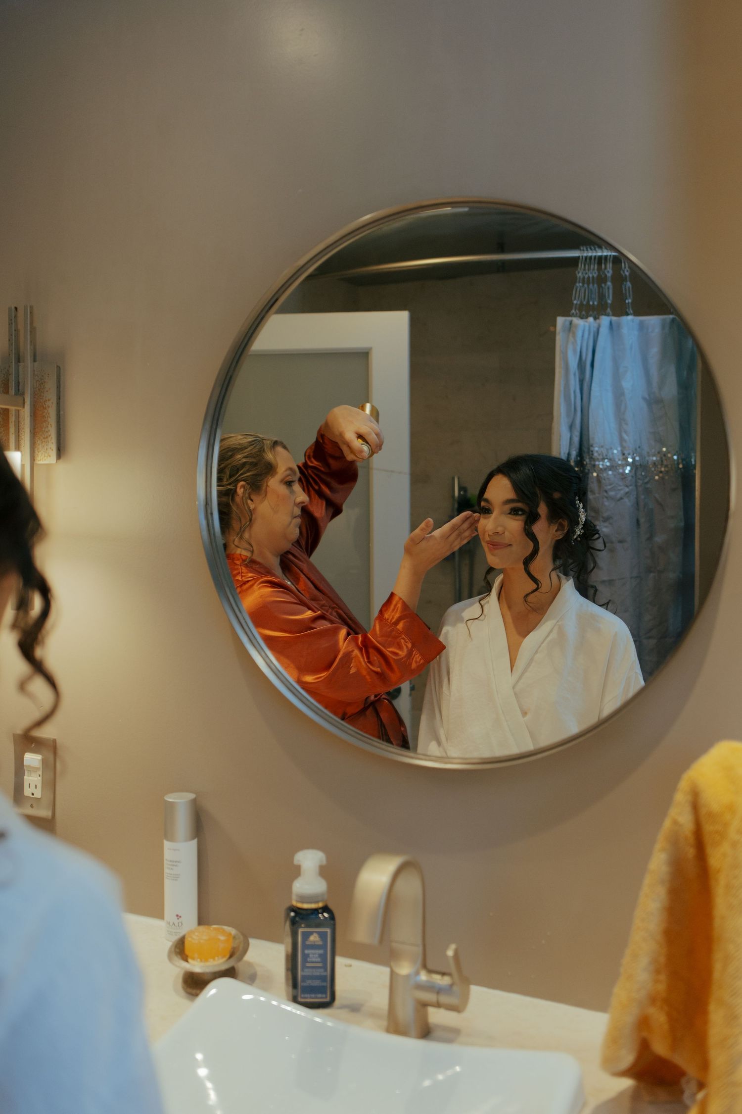 Bride getting her makeup done by a stylist, reflected in a round bathroom mirror on her wedding morning.