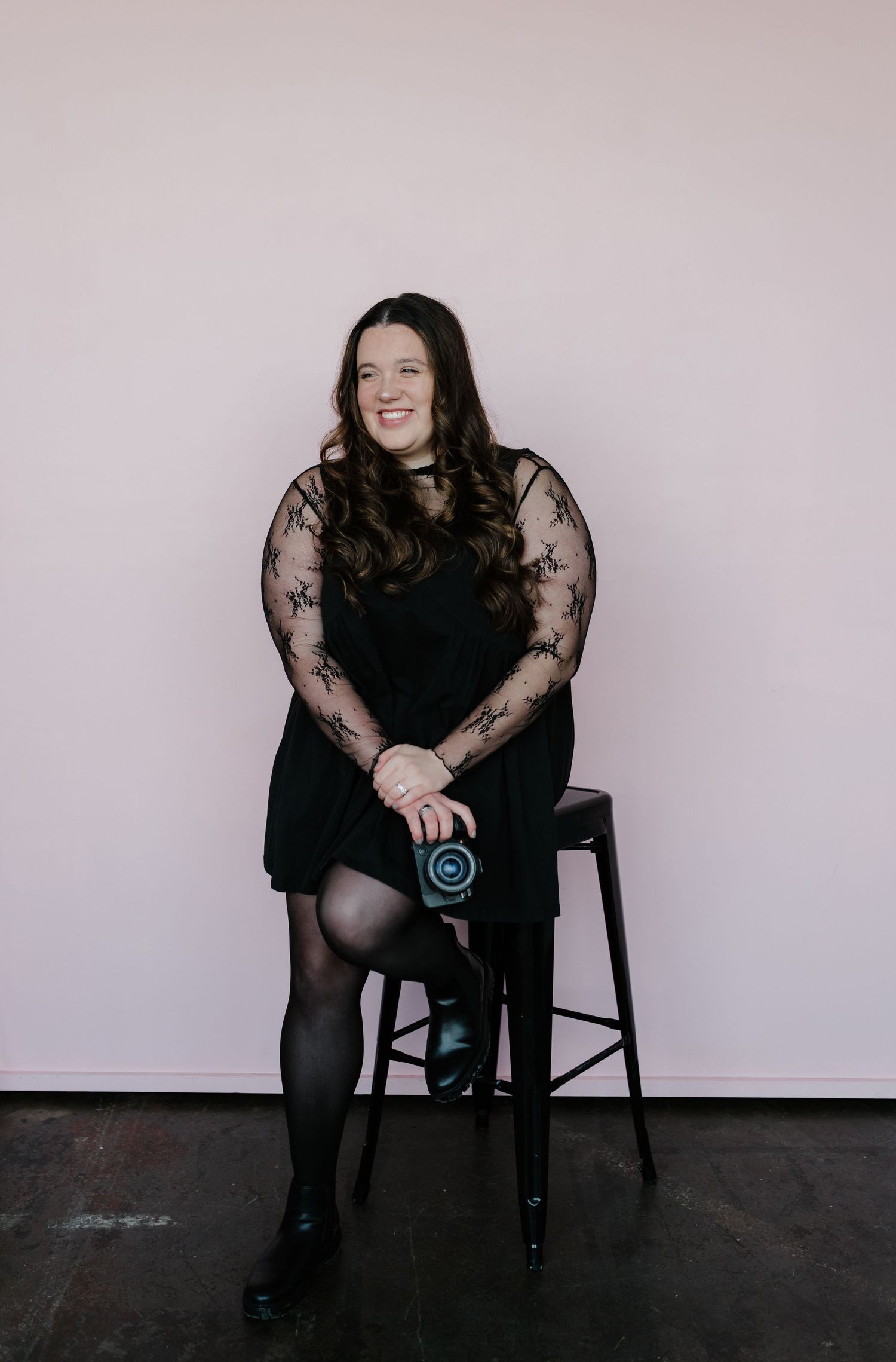 Woman in black lace dress sitting on stool holding camera against white background.