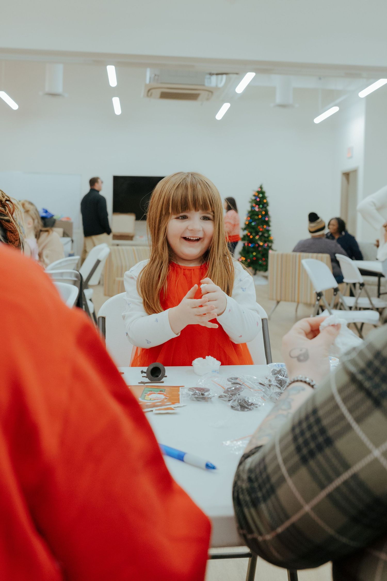 A joyful young girl in a red dress laughs while sitting at a craft table during a Christmas holiday event.