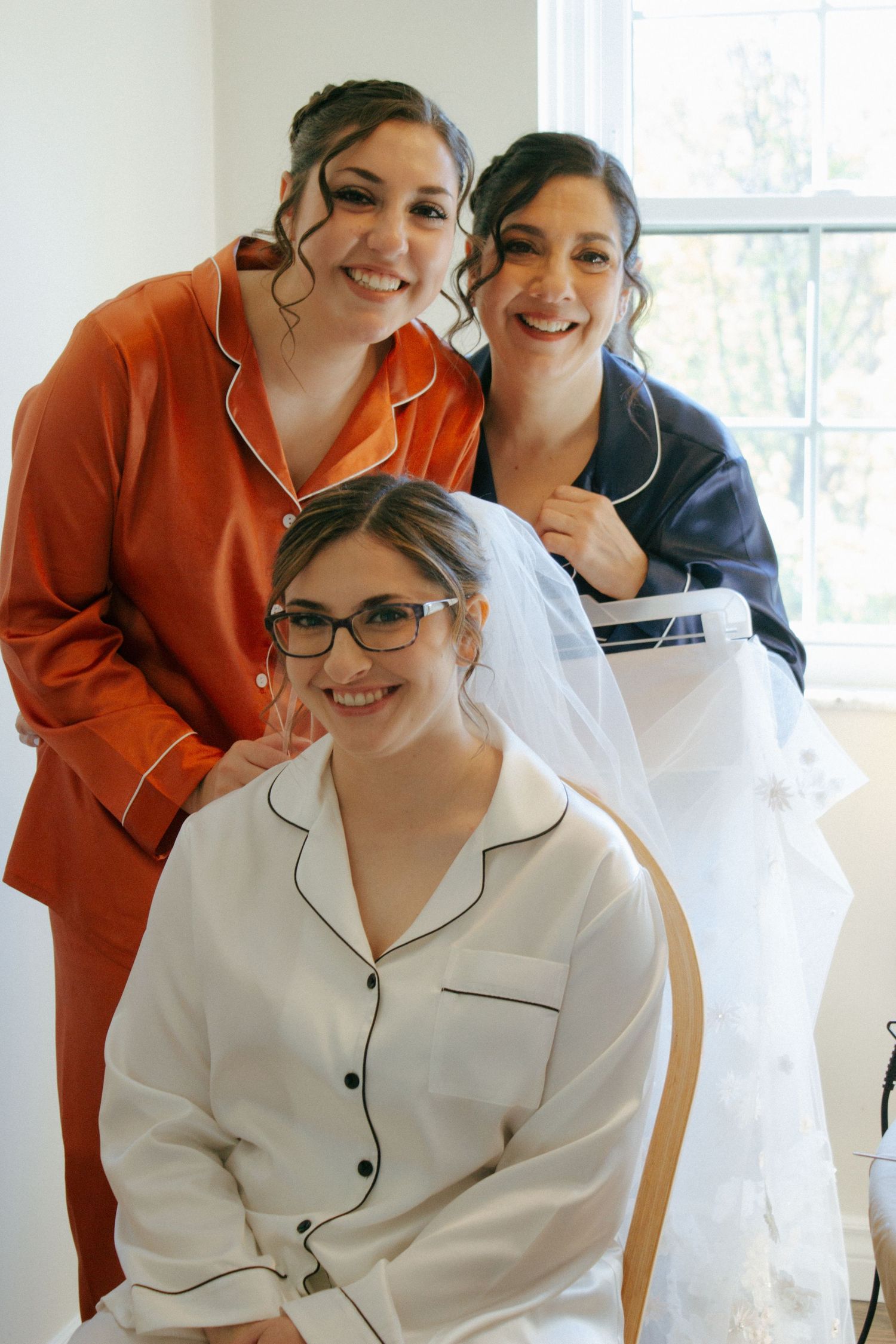 Bride in white pajamas with veil sits with two bridesmaids in colorful satin pajamas before wedding.