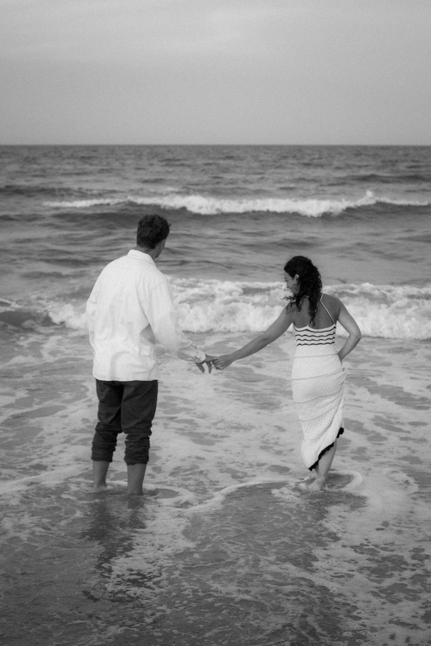 Black and white photo of couple walking hand in hand into ocean waves on a beach.