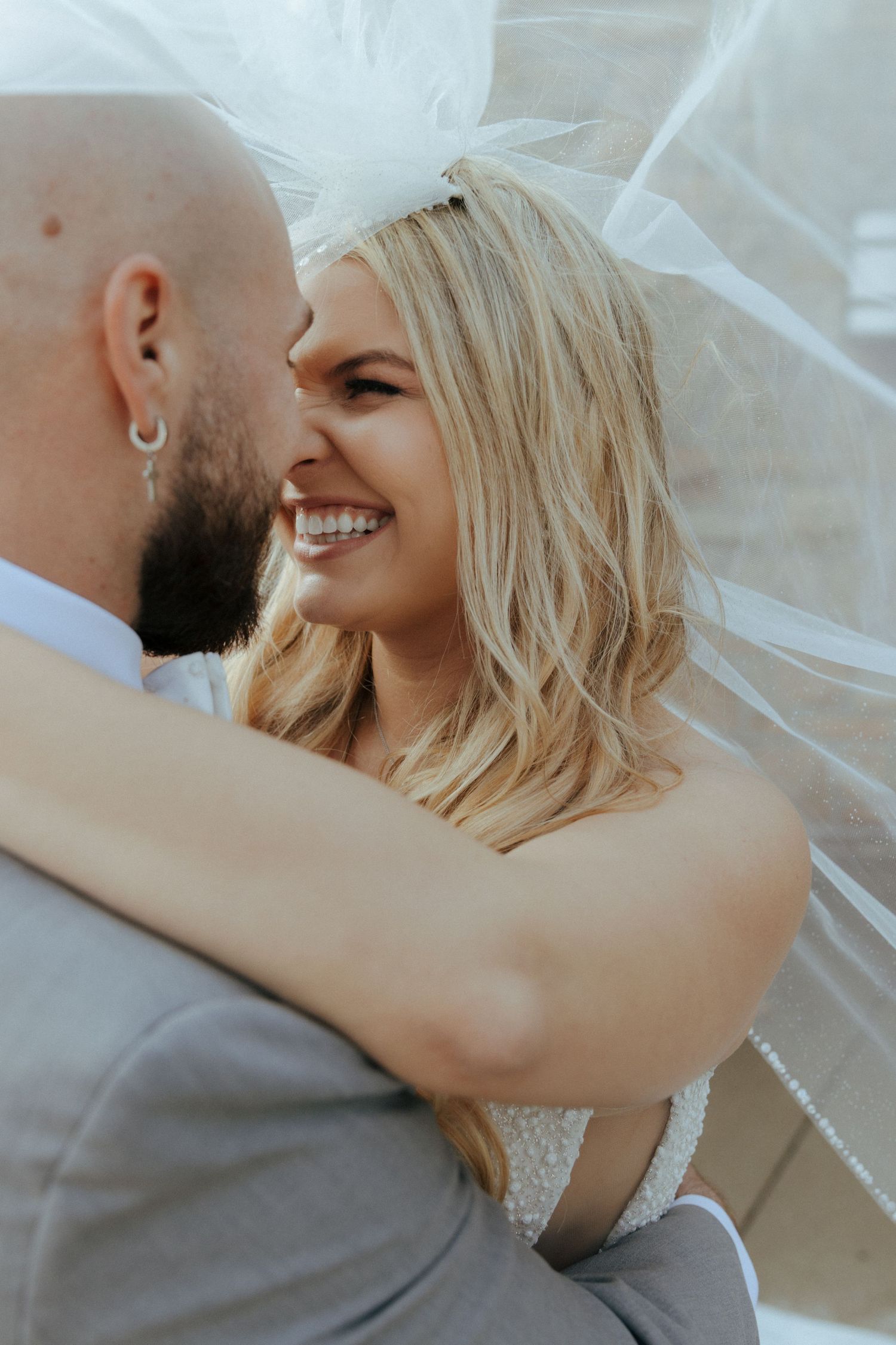 Radiant blonde bride laughing joyfully while embracing her groom beneath her flowing wedding veil.