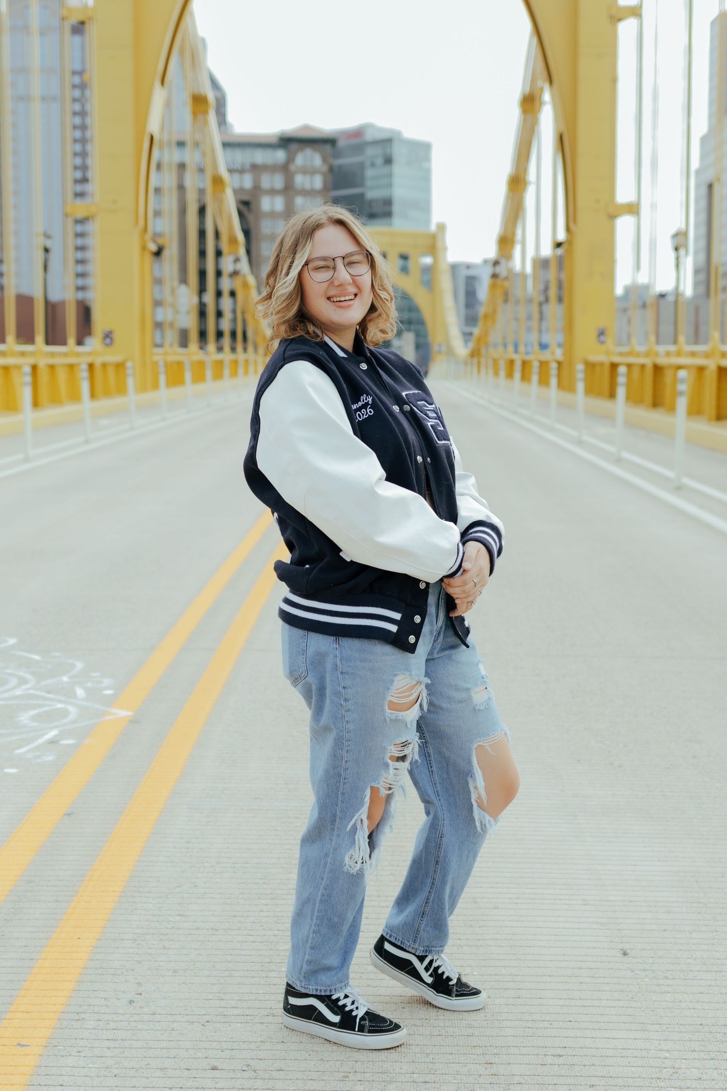 Smiling teen in varsity jacket and ripped jeans poses on yellow Pittsburgh bridge roadway.