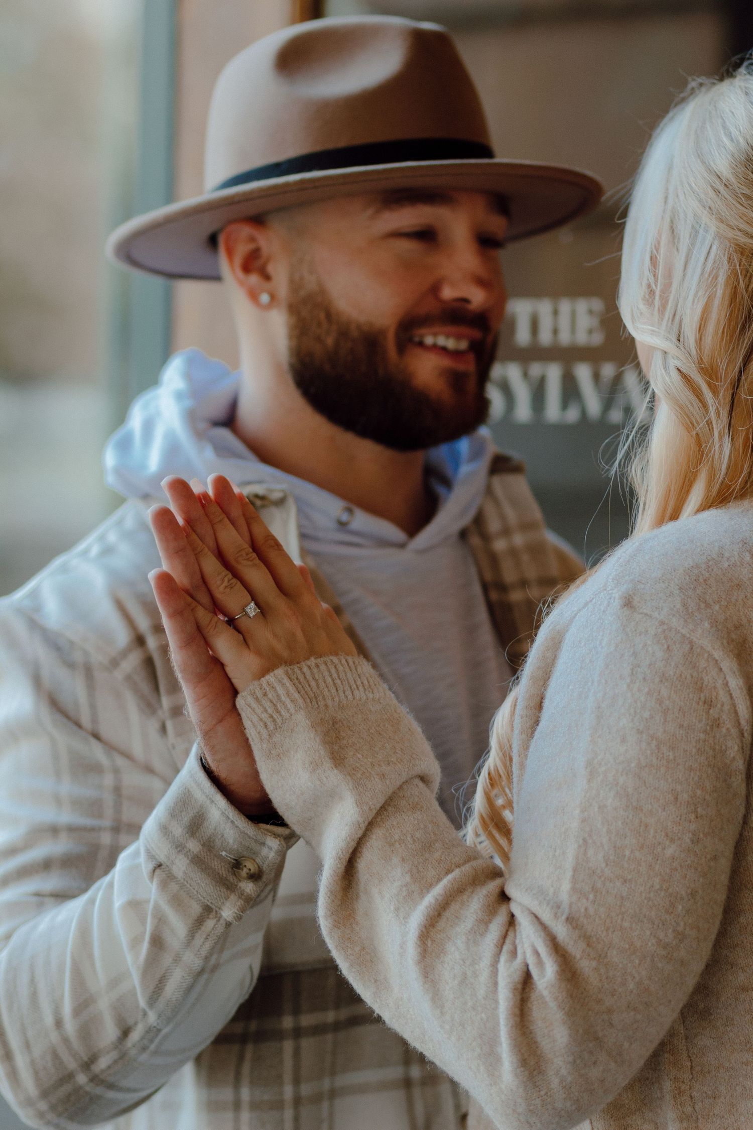 Bearded man in a brown fedora hat smiling at his blonde fiancee while holding hands near a building sign.