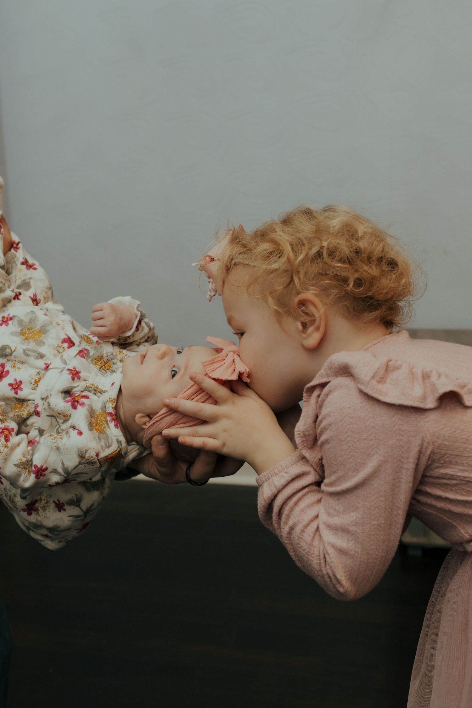 Curly-haired toddler girl in pink dress lovingly kisses a newborn baby held by an adult.