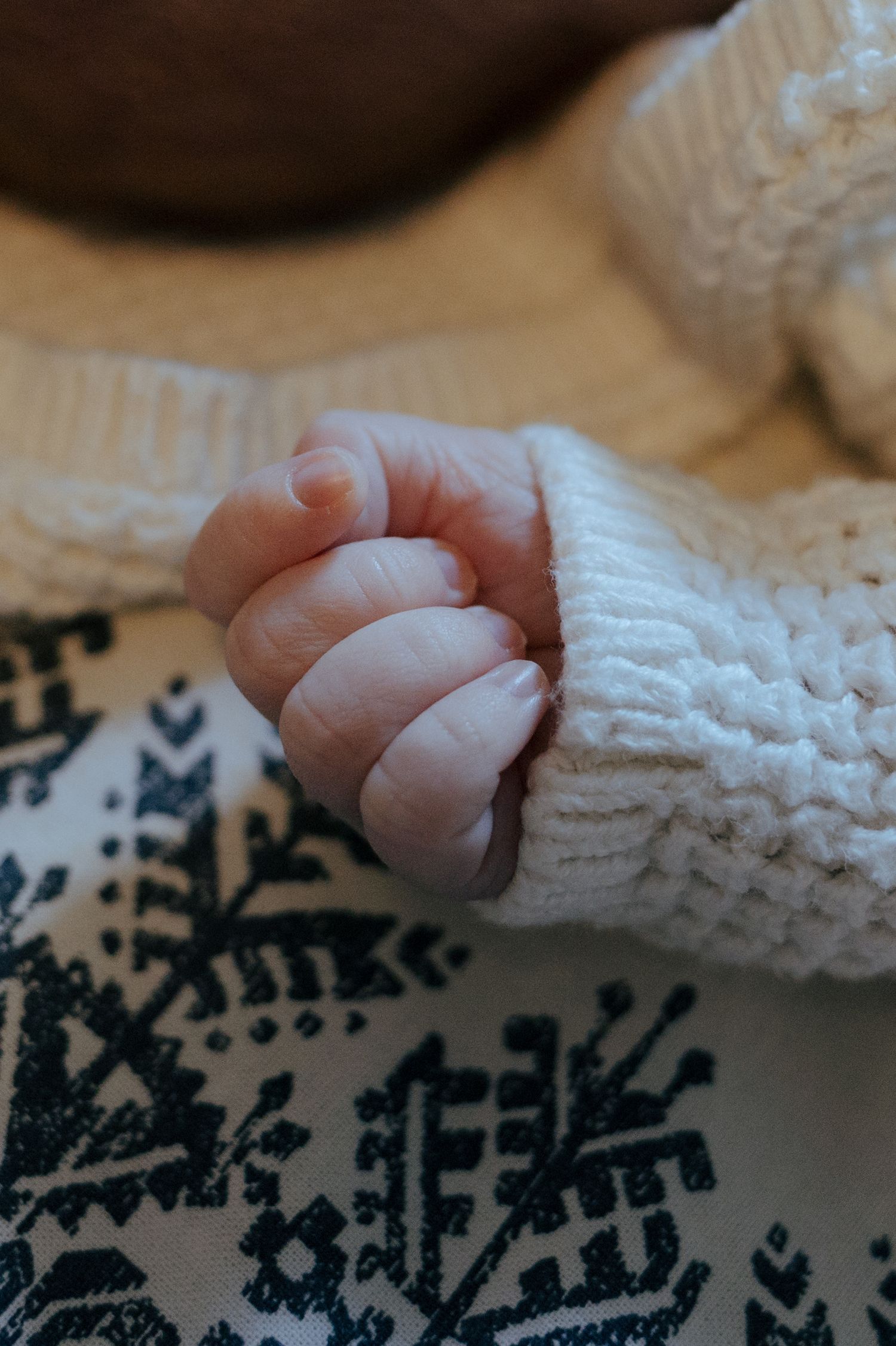 Close-up of a newborn baby's tiny fist in a white knit sweater on patterned blanket.