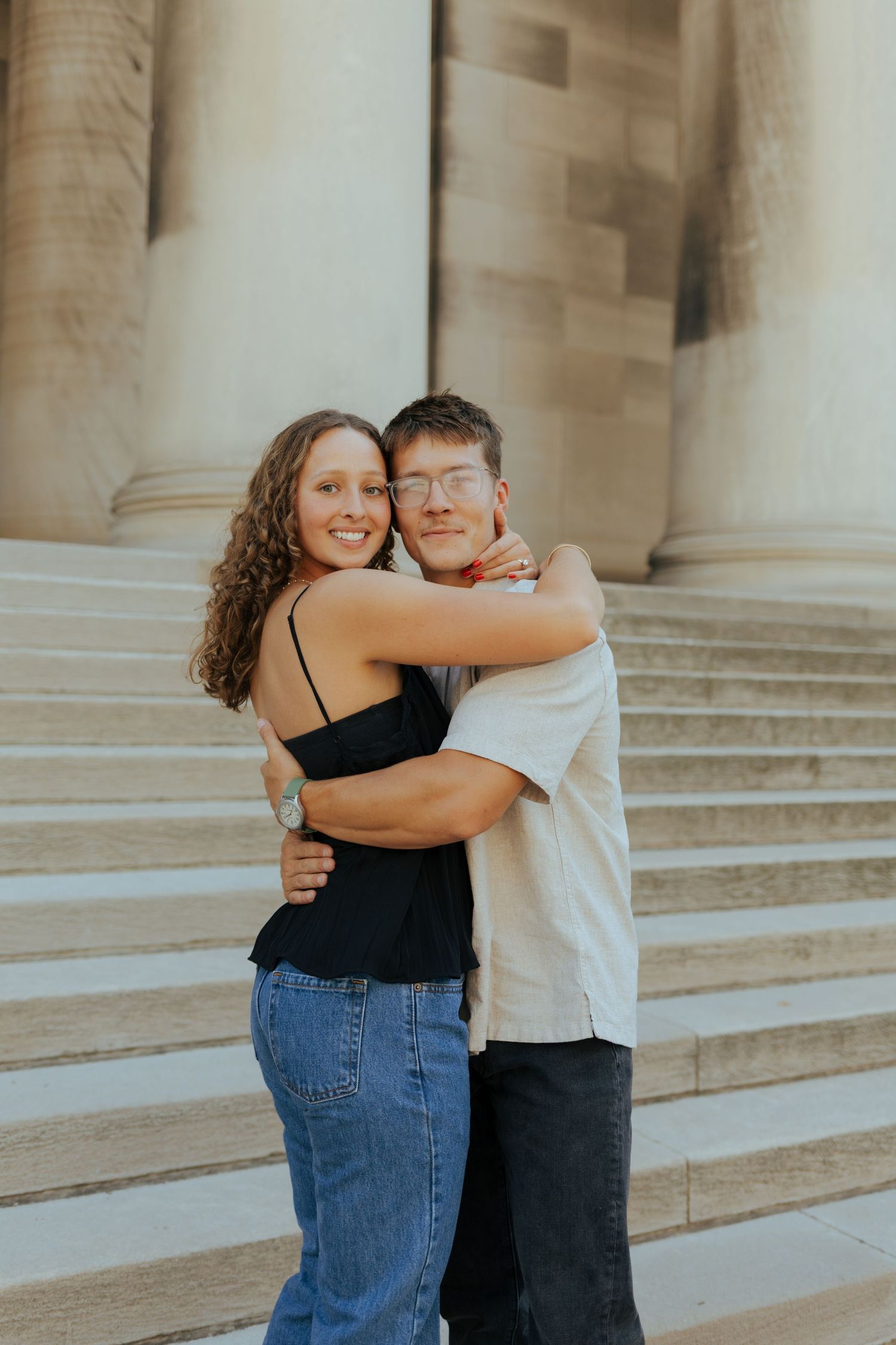 Joyful couple hugging on steps of a grand building with large stone columns behind them.